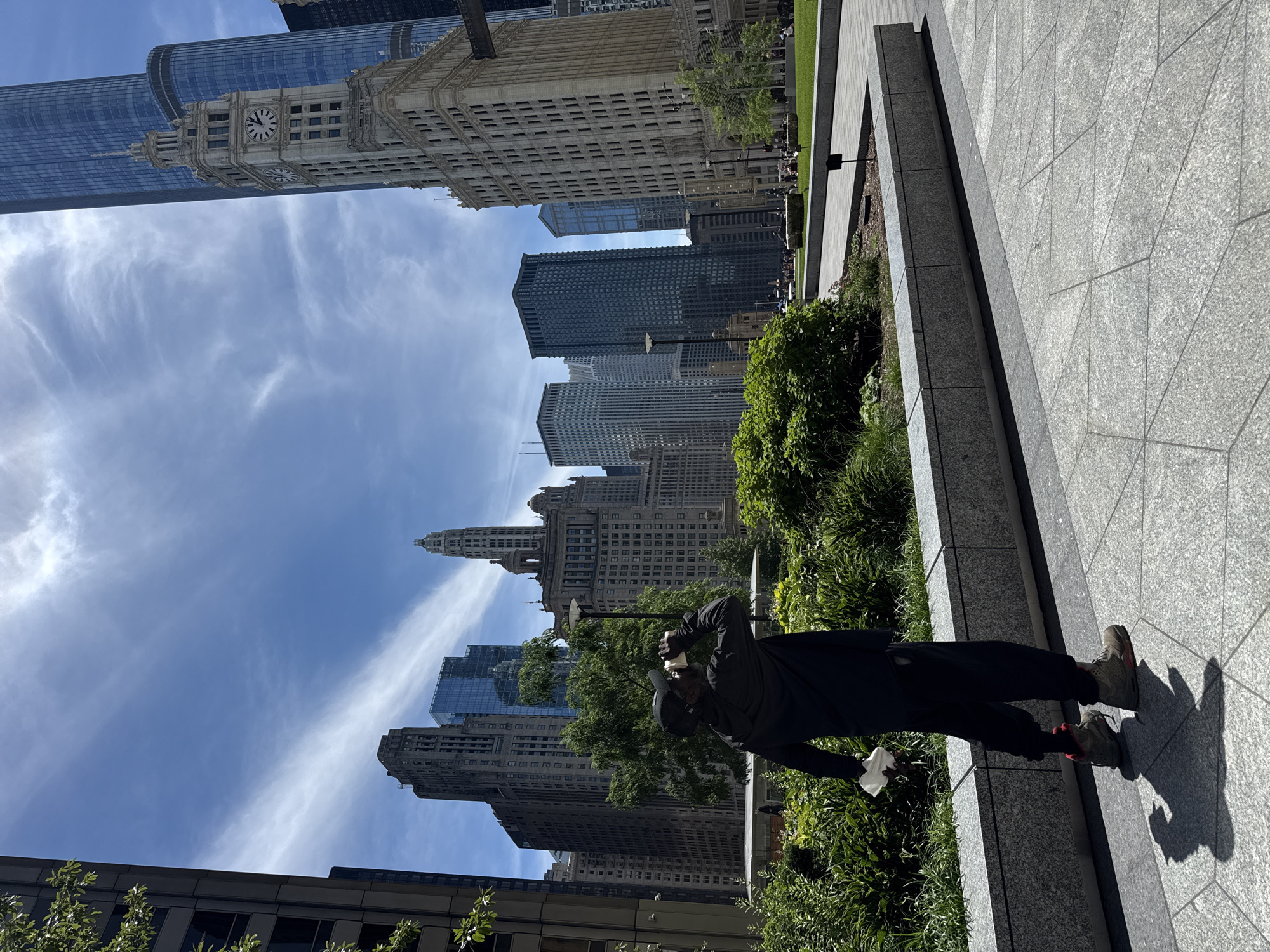 The image depicts a person wearing a black hoodie, black pants, a black baseball cap, and grey and red sneakers, standing in a public plaza in Chicago, Illinois, United States of America. The person is seen holding a white disposable cup to their lips, seemingly drinking. They are also holding a white tissue in their left hand. The plaza features a manicured garden with lush green bushes and trees in the foreground. Behind the garden, a pathway made of grey square tiles is visible, leading towards a grassy area where several people are seen relaxing. In the background, the iconic Chicago skyline dominates the scene, with tall skyscrapers of various architectural styles, including the historic Wrigley Building with its prominent clock tower. The sky is bright blue with scattered white clouds, indicating a clear day. The image is taken from a slightly elevated angle, looking towards the north or northeast.