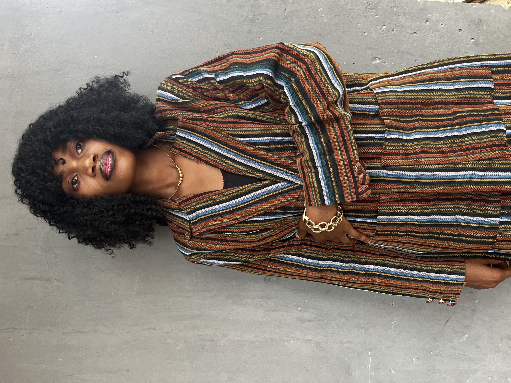 An African woman with dark skin, wearing a multi-colored striped suit, lies on a gray concrete surface. Her suit features horizontal stripes in hues of orange, black, white, and blue. She wears a gold chain necklace and a chunky gold bracelet on her wrist. Her dark curly hair frames her face. The location is indicated as Badagry, Nigeria.