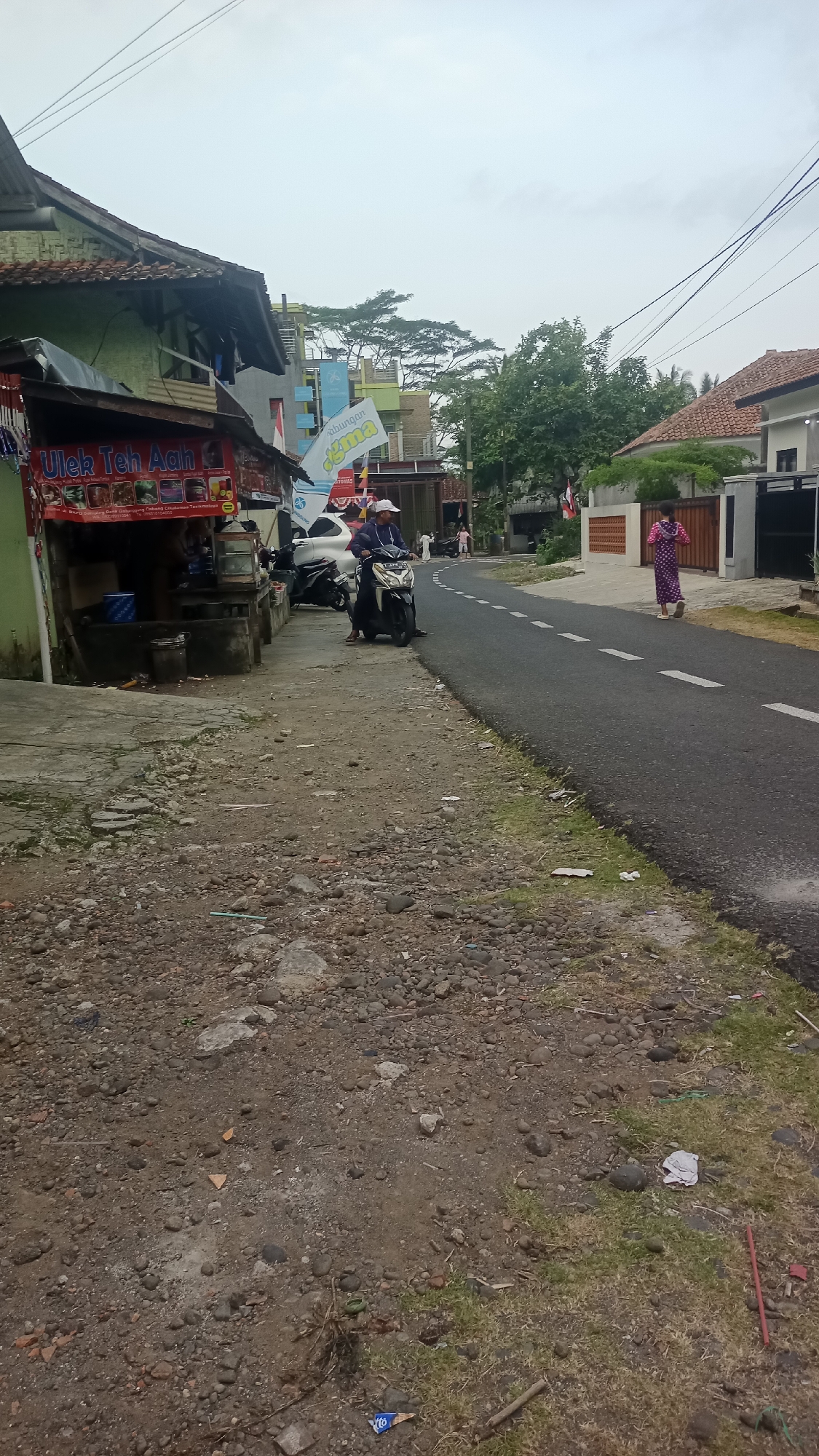 The image depicts a street scene in the city of Pakemitan, Indonesia, under an overcast sky. On the left side of the road, a small food stall with a green facade displays a banner that reads "Ulek Teh Aah". Several motorcycles are parked near the stall. A person wearing a white cap and a dark jacket is seated on a motorcycle, facing forward.
The road itself is paved and has white dashed lines marking the lanes. A woman in a long, patterned dress walks away from the viewer on the right side of the road. Further down the road, there are several buildings and trees. The ground along the side of the road is unpaved, covered with gravel and some sparse vegetation. Litter is visible on the ground.
