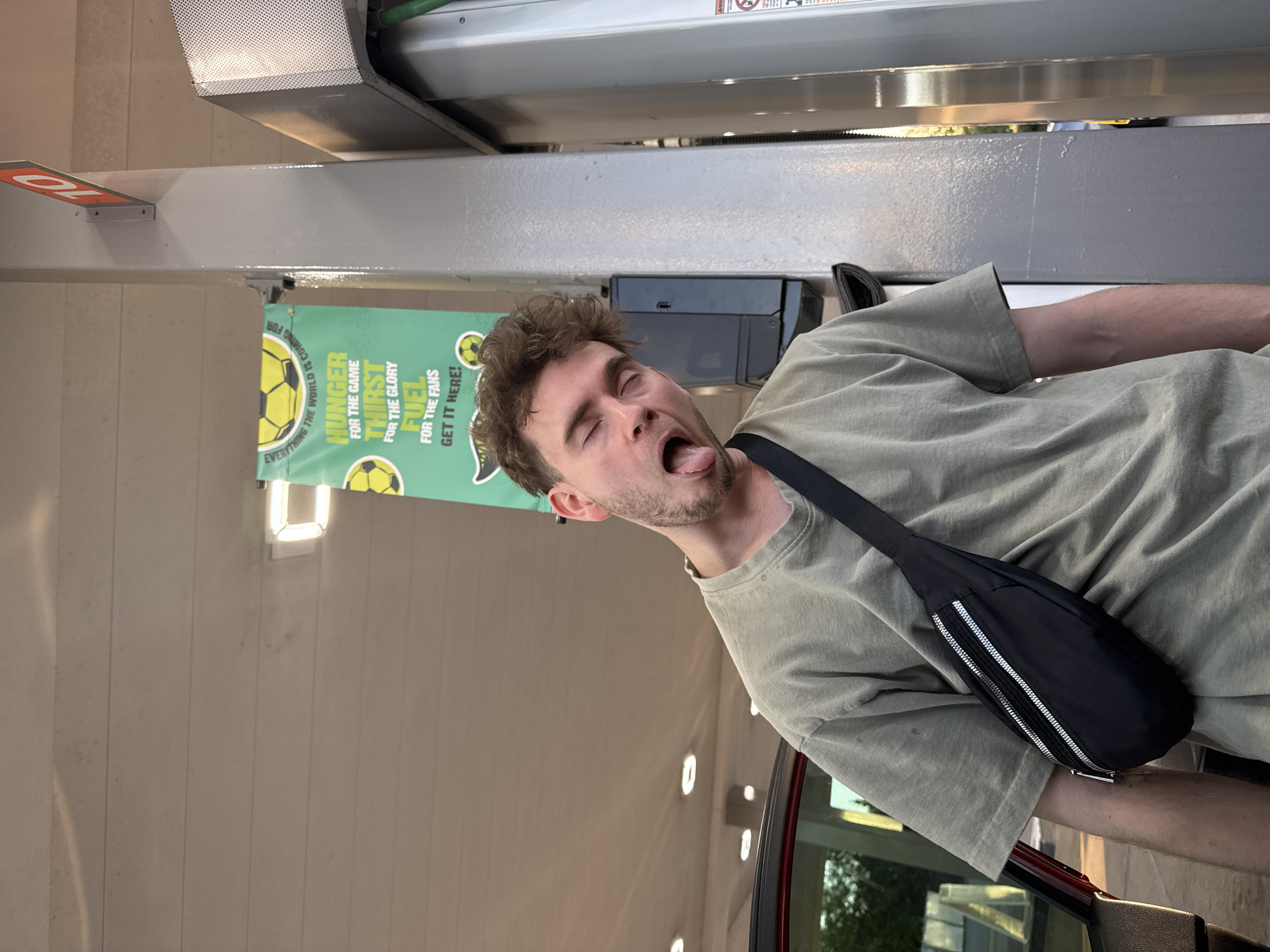 A young man is photographed at a gas station in Fife, Washington, with his eyes closed and his tongue sticking out, as if he is posing for a playful photo. He is wearing a light green t-shirt and a black fanny pack. Behind him, a green banner with yellow soccer balls and text about "Hunger for the game, thirst for the glory, fuel for the fans" is visible. The man is leaning against a gray support column, and a gas pump is to his right. The background shows the ceiling of the gas station canopy and part of a red car.