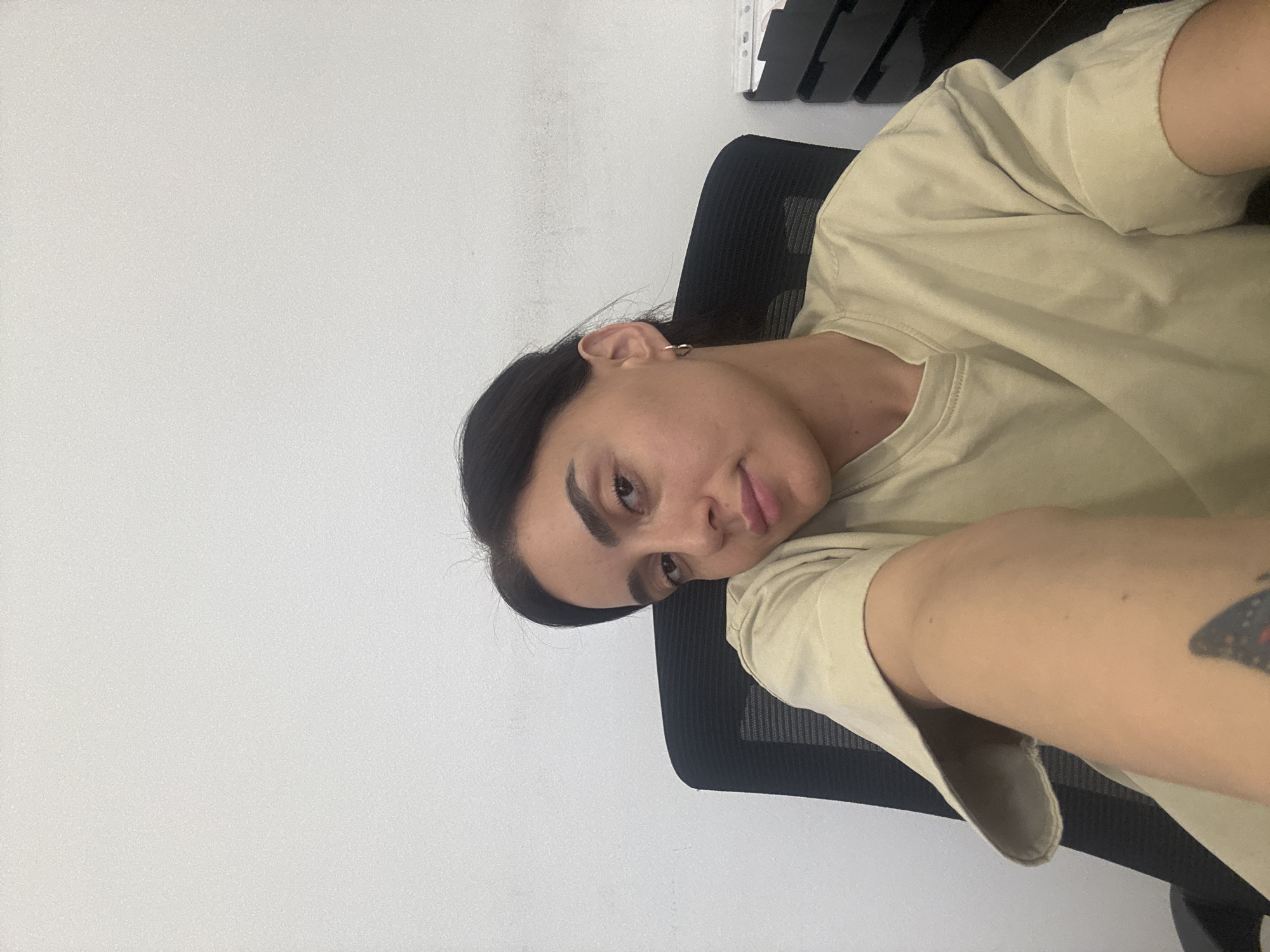 A young woman with dark hair and a tan complexion is seated in an office chair, with her body angled towards the camera. She is wearing a light beige t-shirt and a small hoop earring in her left ear. Her right arm is extended forward, and a butterfly tattoo is visible on her forearm. The background consists of a plain white wall. To the right of the woman, a black office chair and a filing cabinet are partially visible. The overall impression is of a casual, indoor setting, possibly an office, given the presence of the chair and filing cabinet. The image does not contain any specific indicators of its location within Tbilisi, Georgia.