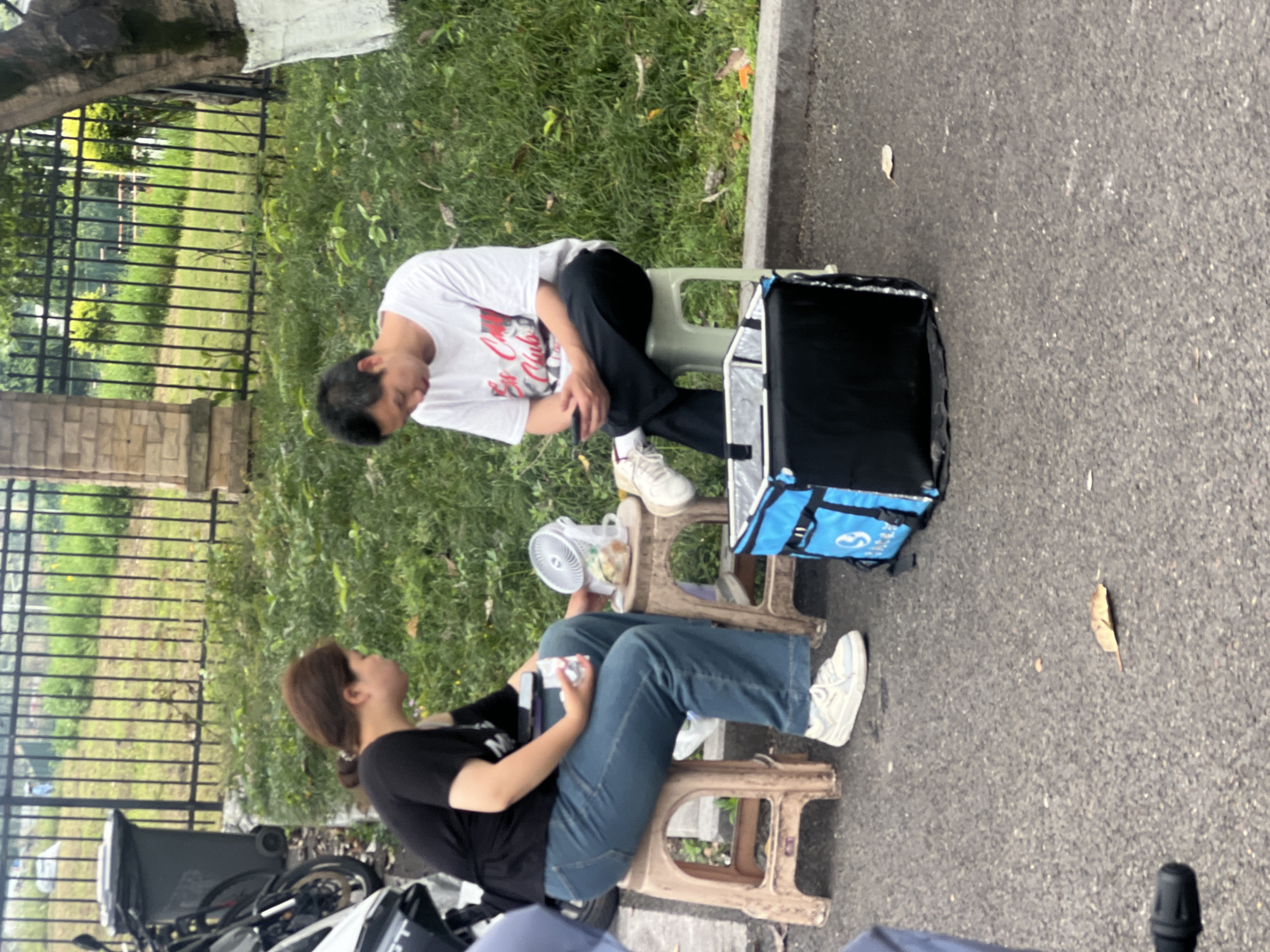 In Jiangjin, China, two individuals are seated outdoors. A man in a white t-shirt and black pants sits on a small green stool. He appears to be focused on his hands. Adjacent to him is a large black and blue insulated food delivery bag. A woman in a black t-shirt and blue jeans is seated on a similar small beige stool, holding a phone or small electronic device. A small white electric fan is placed between them. A motorcycle is parked nearby. A fence and grass border the scene, while a paved road fills the remaining space.