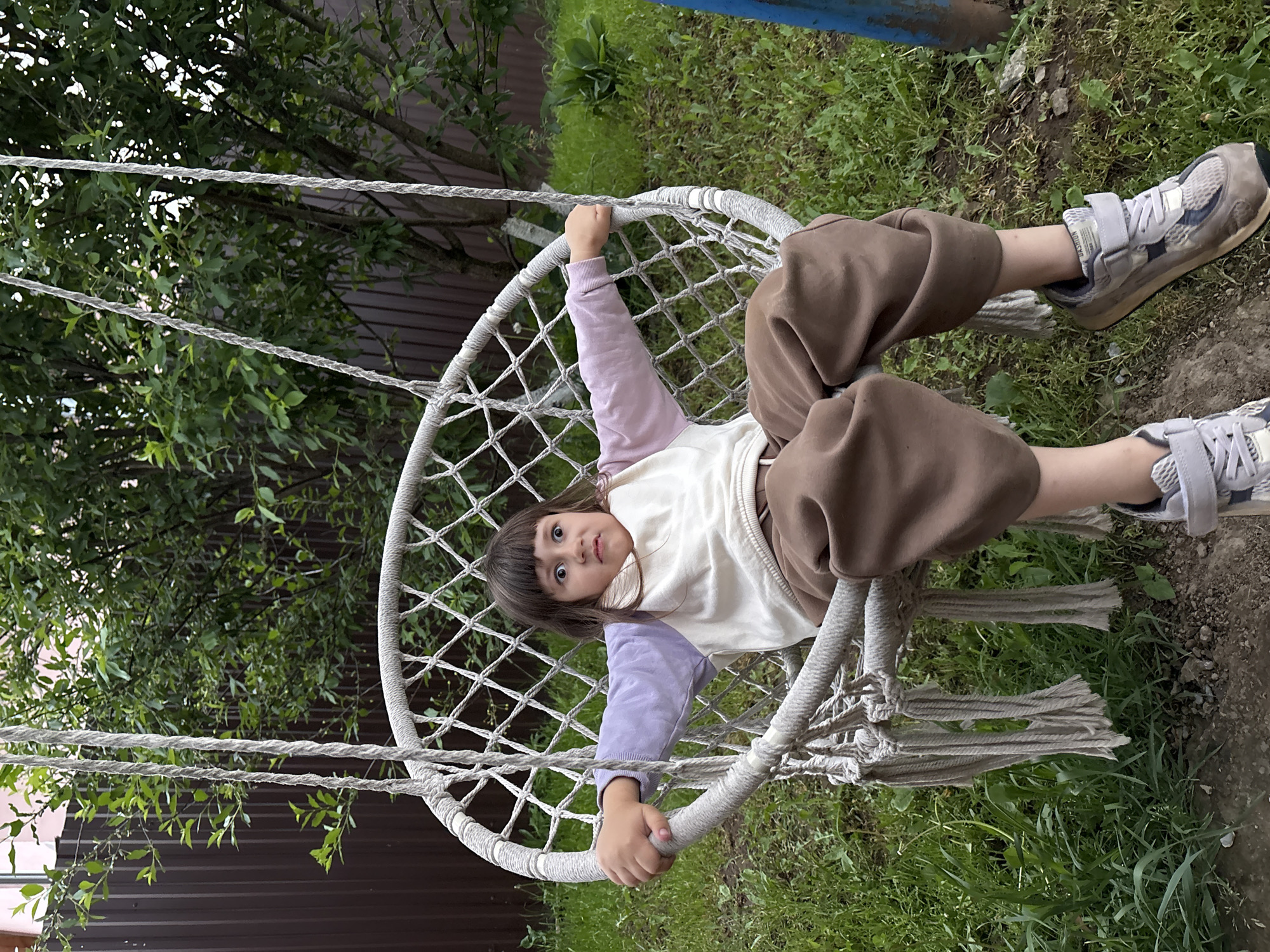 A young girl sits in a white rope swing outdoors, likely in a backyard or garden setting. She is wearing a long-sleeved shirt with a lavender sleeve and a white center, brown pants, and grey sneakers. The swing is suspended from ropes, and the background shows a fence, trees, and grass. The location is Прикамский, Russia.