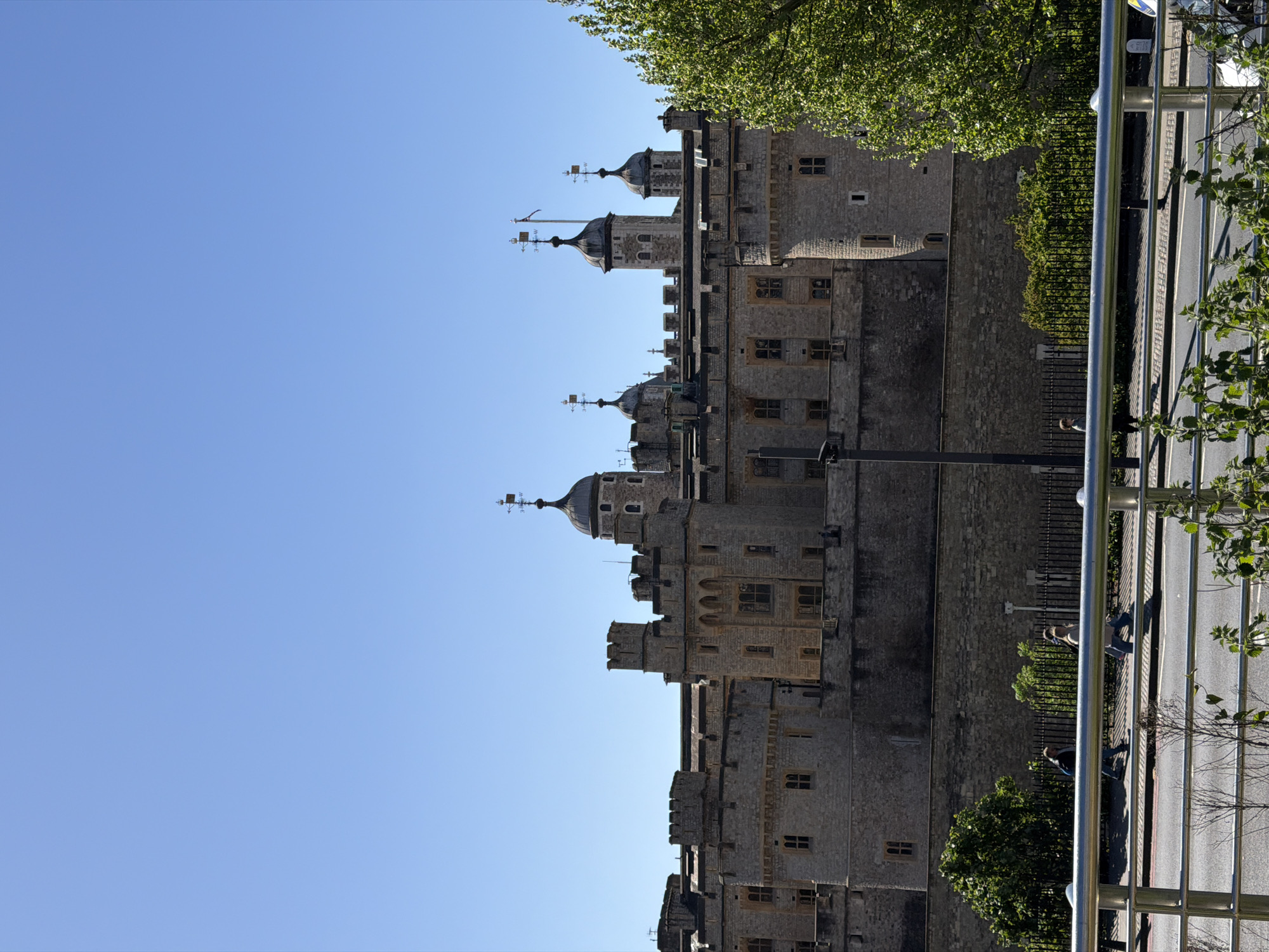A bright, daytime photograph captures a section of the historic Tower of London under a clear blue sky. The imposing stone structure dominates the scene, featuring multiple grey turrets with dark, domed roofs, many topped with finials and weather vanes. The castle walls are constructed from light-colored stone blocks, punctuated by numerous small, rectangular, and arched windows. Sections of battlements are visible along the tops of the walls.
In the foreground, running parallel to the castle, is a street or road. A modern-style silver metal railing acts as a barrier, separating the castle grounds from the public thoroughfare. Several indistinct figures of people are visible on the sidewalk adjacent to the road, appearing to be walking. Green foliage from trees is scattered throughout the frame; some tree branches overhang the castle from the upper right, while other trees are situated closer to the street level.
The bright sunlight indicates a clear day, likely mid-morning to late afternoon, with noticeable shadows. The scene depicts a typical day around the iconic London landmark, with people moving along the public path. No discernible text is visible anywhere in the image.