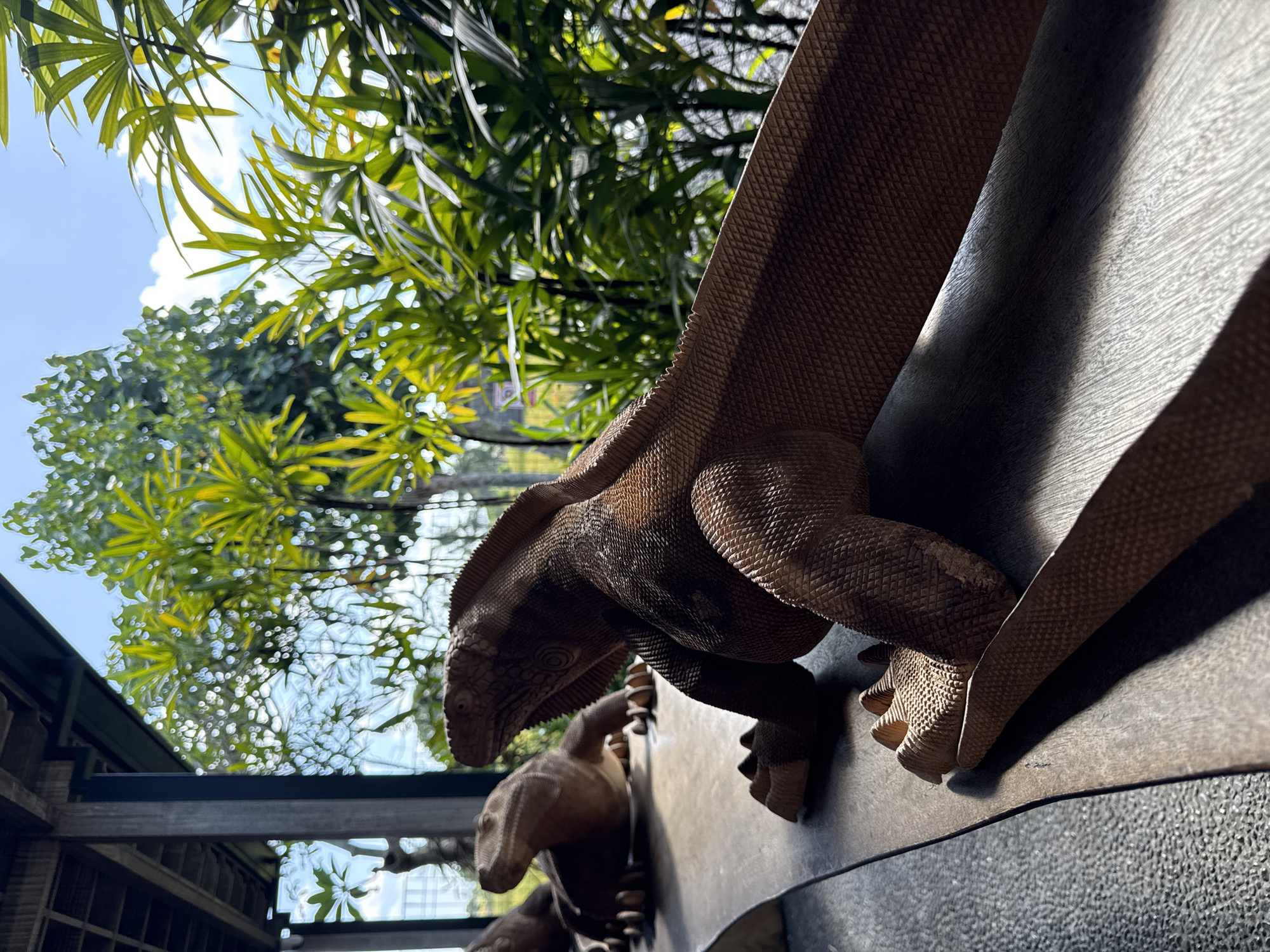 The image shows a wooden carving of a lizard resting on a wooden surface, with another lizard carving in the background. Lush green foliage and a bright blue sky are visible behind the carvings, suggesting an outdoor setting. The lighting indicates it is daytime. The scene is likely located in Sukawati, Indonesia, a region known for its art markets and wood carvings.