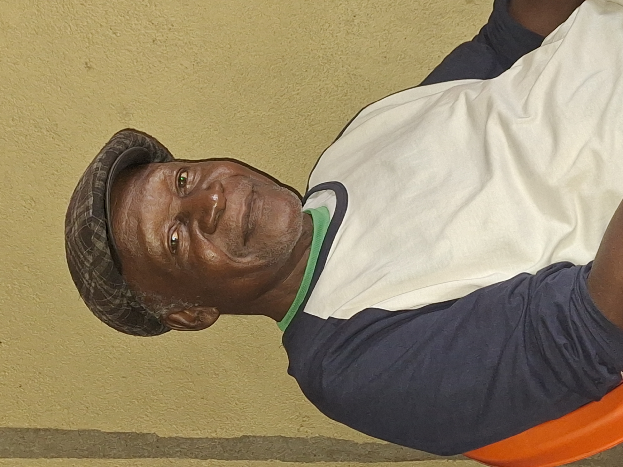 This is a rotated portrait image showing a dark-skinned man, likely in Sabon Tasha, Nigeria. His head is positioned towards the right side of the frame, looking directly at the viewer with a calm, neutral expression. His eyes are dark brown with visible reflections.
The man is wearing a dark, plaid-patterned flat cap. He is dressed in a two-tone t-shirt featuring a white main body, dark blue sleeves, and a bright green trim around the collar. A portion of dark blue clothing, possibly shorts or trousers, is visible below the t-shirt. He appears to be seated on an orange object, likely a chair or stool, with only a small segment visible at the bottom right.
The background consists of a plain, light yellowish-brown or tan wall with a subtle textured finish. The lighting appears even, suggesting daytime conditions, possibly indoors or in a shaded outdoor setting. There is no visible text in the image.