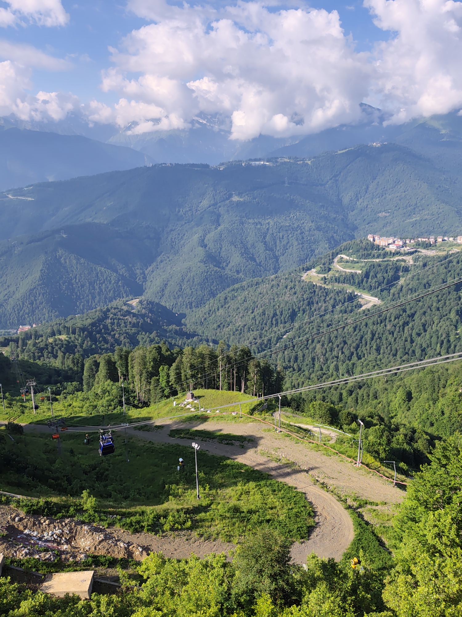The image captures a panoramic view of a mountainous landscape in Estosadok, Russia, likely during daytime under a partly cloudy sky. In the foreground, a ski lift with multiple gondolas is visible, traversing cables strung between support towers. The terrain consists of steep, green slopes covered in dense trees and a dirt path winding its way down. Some scattered construction materials, including concrete and rocks, are present on the left side of the foreground. In the mid-ground, more ski lift infrastructure, including towers and cables, is evident, along with a paved road snaking through the forested mountainside. A cluster of buildings, appearing to be a resort or residential complex, is situated on a distant mountain ridge. The background is dominated by layers of lush green mountains, receding into the hazy distance, with patches of white snow visible on the highest peaks. The overall scene suggests a summer or early autumn view of a mountain resort area.