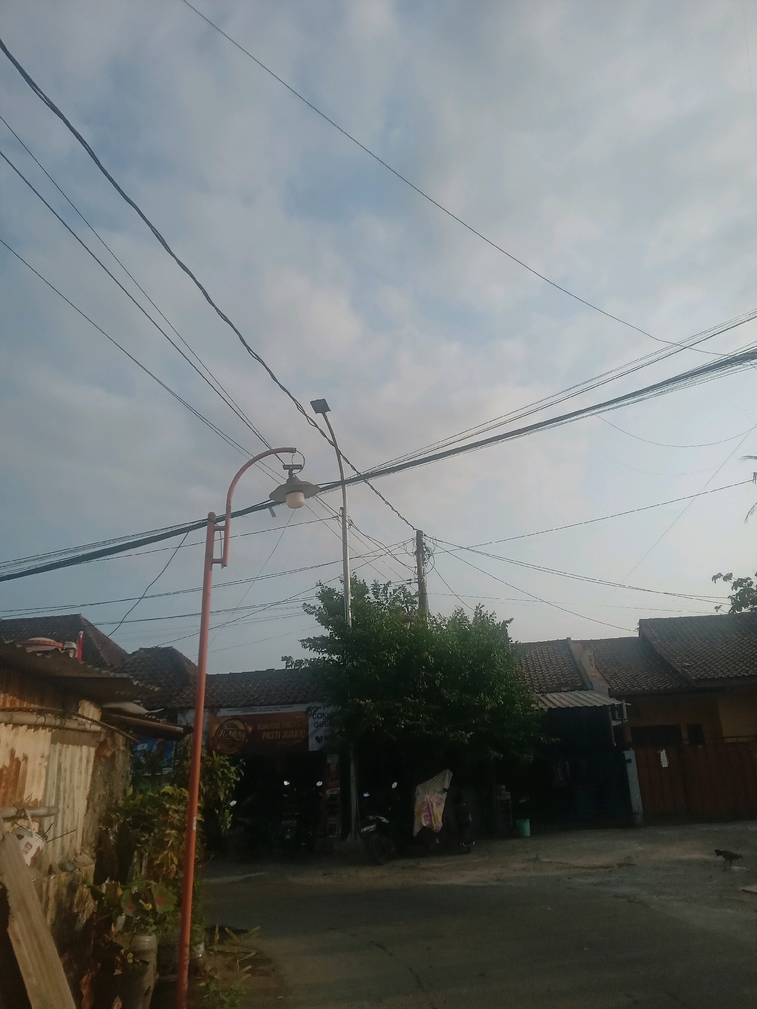 The image depicts a street scene in Banguntapan, Indonesia. The sky is cloudy and overcast, with numerous electrical wires strung across the frame. In the foreground on the left, a corrugated metal structure stands next to a red lamppost. Behind the lamppost, a dense green tree obscures the view of the buildings and a street. The buildings are single-story structures with tiled roofs, suggesting a residential or commercial area. A sign is visible above an open doorway, possibly indicating a business. A few motorcycles are parked near the buildings, and a small, dark animal, possibly a cat or dog, is walking on the road in the middle ground on the right. The road surface appears to be asphalt. The overall impression is of a typical urban or suburban street in Indonesia, with evidence of infrastructure and daily life.