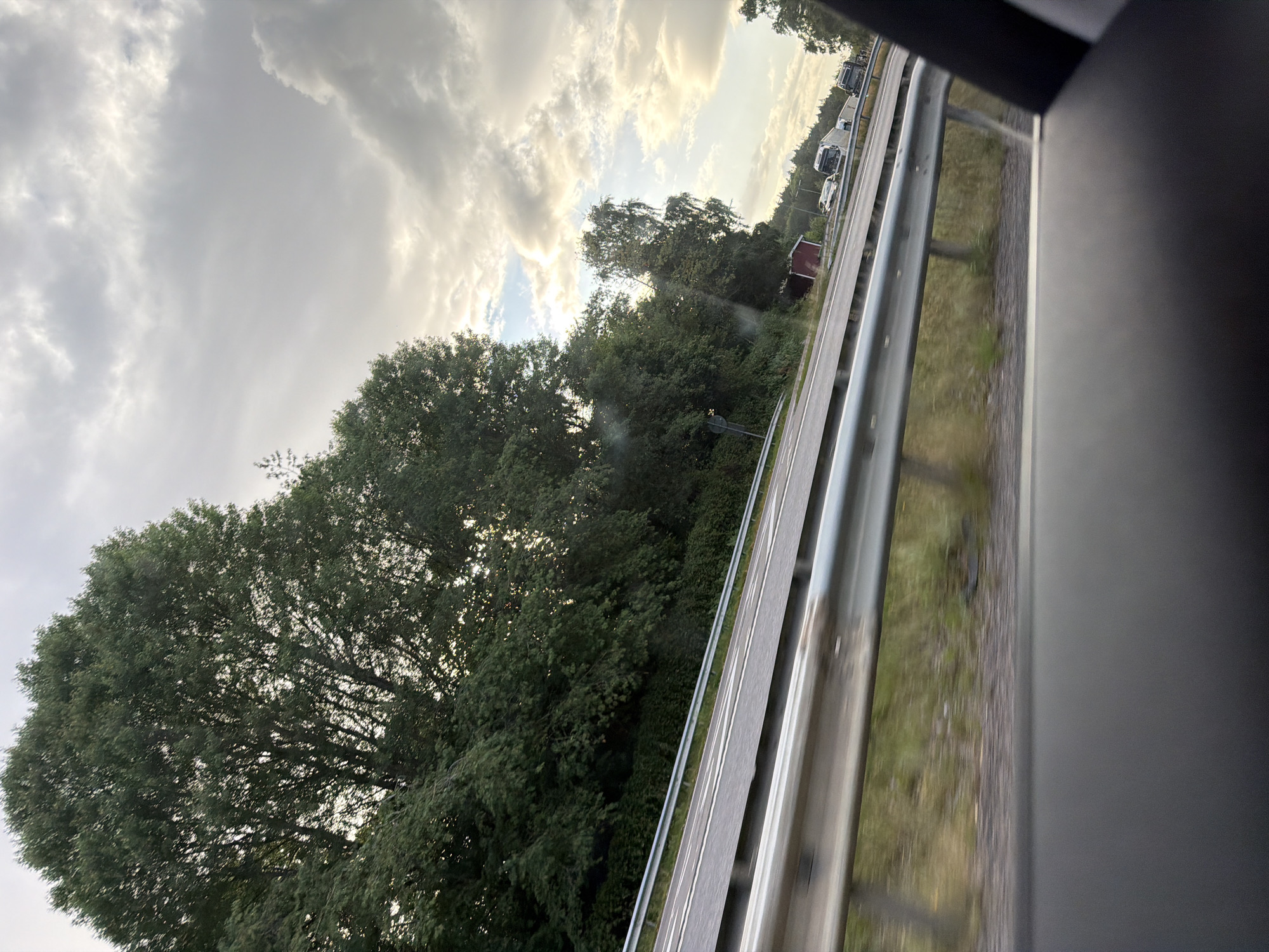 The image depicts a view from inside a vehicle looking out towards a highway. On the left side of the highway, there is a dense line of tall, green trees. The highway itself has two lanes visible, separated by white lines, and is bordered by a silver metal guardrail in the foreground and middle ground. Beyond the highway, the terrain is covered with green grass and some dry patches. In the distance, on the right side of the highway, several vehicles are visible, including trucks and cars. The sky is filled with clouds, with patches of sunlight breaking through, suggesting either early morning or late afternoon. A small red building is also visible near the highway. The scene is located on a highway in or near Frillesås, Sweden.