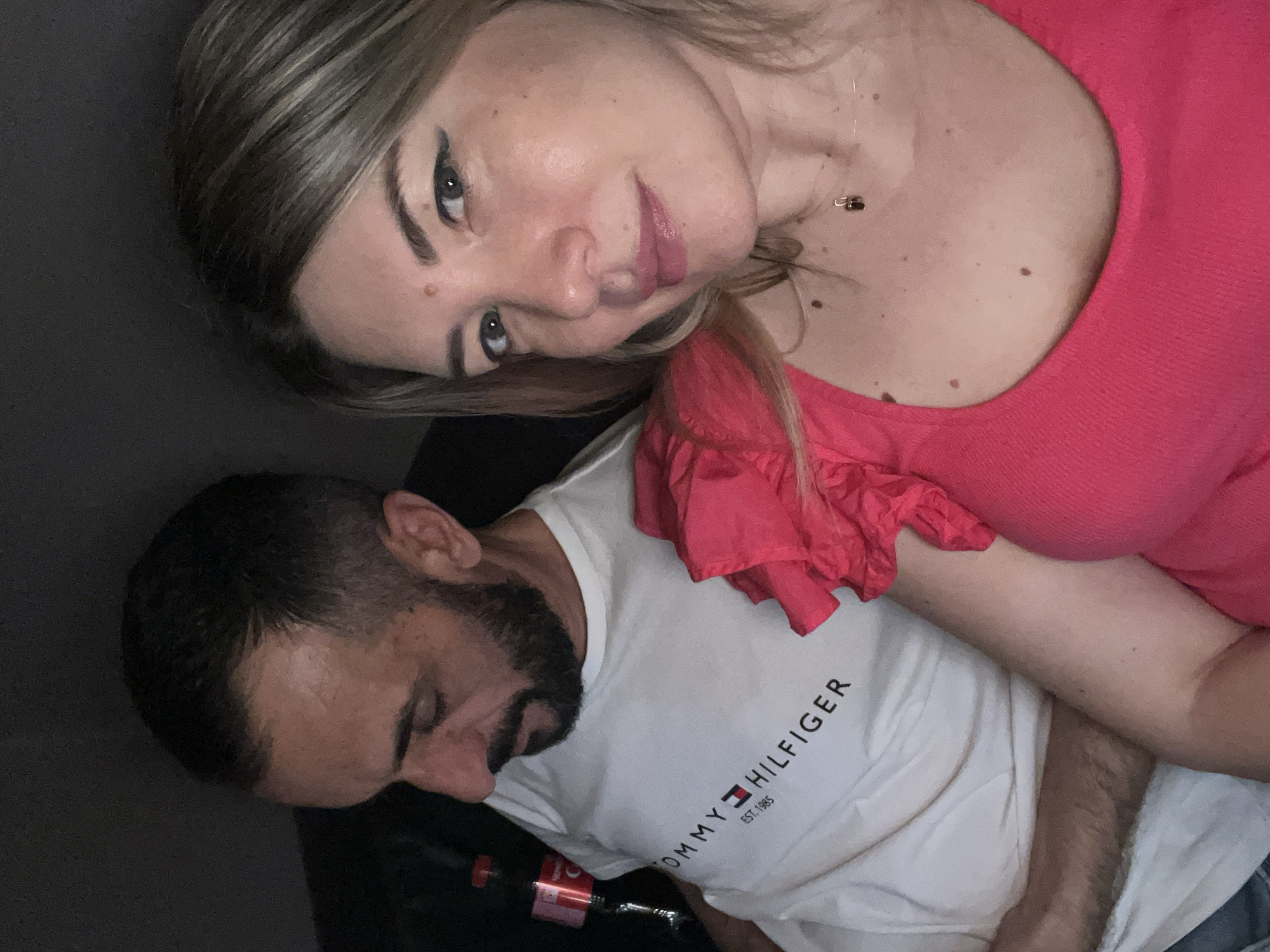 The image, captured in an indoor, low-light setting in the city of Sochi, Russia, features two individuals. In the foreground, positioned on the right, is a light-skinned female with shoulder-length, light-brown hair. She wears a vibrant fuchsia-colored top with ruffled detailing on the right shoulder and a delicate gold-colored chain necklace bearing a small, dark (possibly lock-shaped) pendant. Her face, directly illuminated, shows light pink lipstick, dark winged eyeliner, and noticeable moles, including one on her upper forehead and several on her décolletage. She looks directly into the camera with a slight, closed-lip smile.
To the left and slightly behind the female is a male with olive skin, short dark hair, and a full dark beard and mustache. He wears a white t-shirt featuring the black 'TOMMY HILFIGER' logo and 'EST. 1985' beneath it. His head is turned downwards, and his gaze is directed away from the camera. Partially visible behind his left shoulder is a red Coca-Cola bottle with a black cap. The background is uniformly dark and indistinct, indicating a dimly lit interior environment. The female's angle and proximity to the camera suggest she is capturing a selfie.