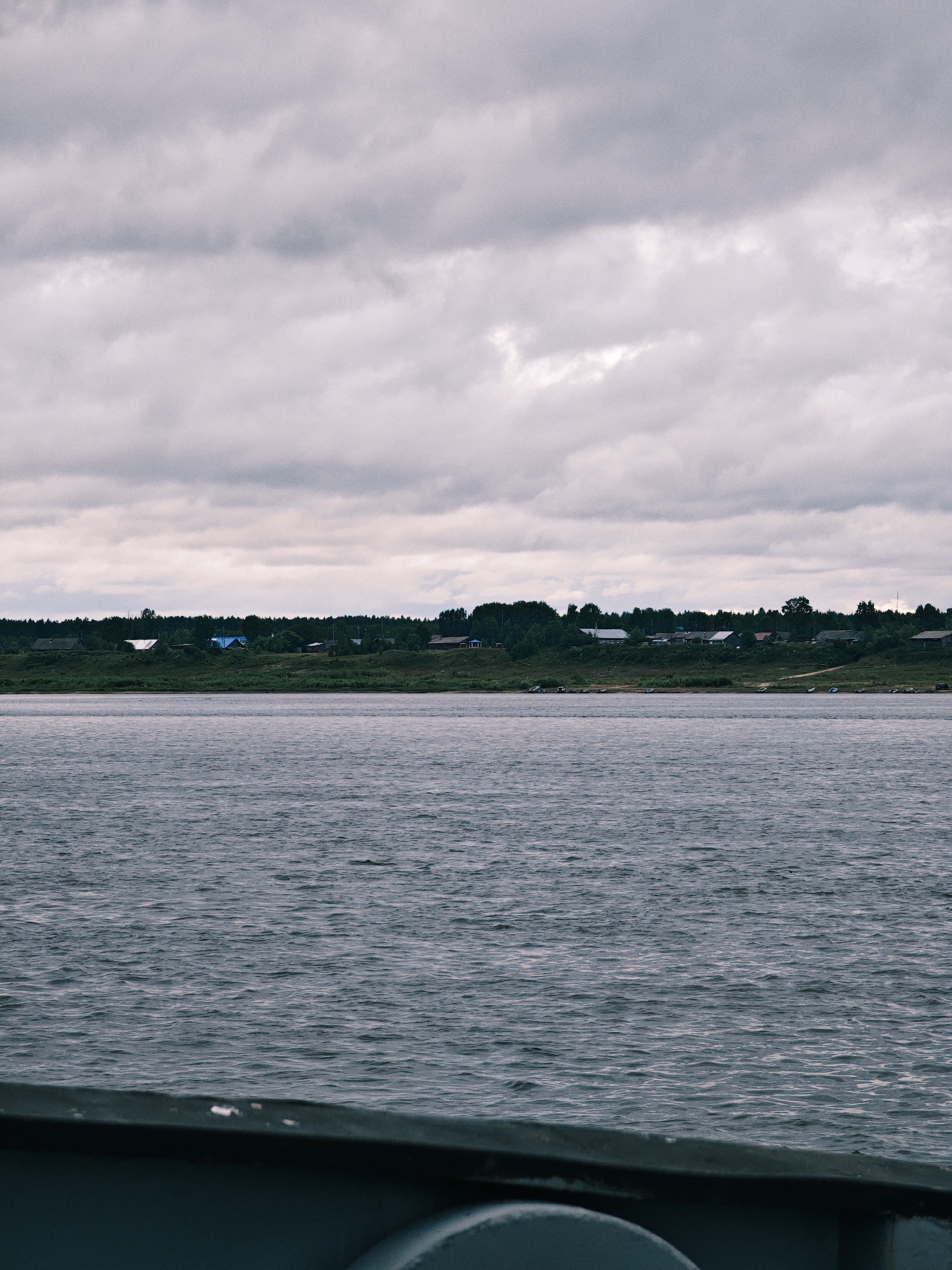 The image shows a wide, choppy river with a village on the far bank. The sky is overcast with heavy, gray clouds. The village consists of several wooden houses with various colored roofs, situated on a grassy embankment lined with trees. Some boats are visible along the shoreline of the village. In the foreground, the dark, blurry edge of what appears to be a vessel is visible, suggesting the photo was taken from a boat on the river. The overall scene is calm and somewhat somber, typical of a cloudy day in a rural setting. The location is a village on the Yenisei River in Zotino, Krasnoyarsk Krai, Russia.
