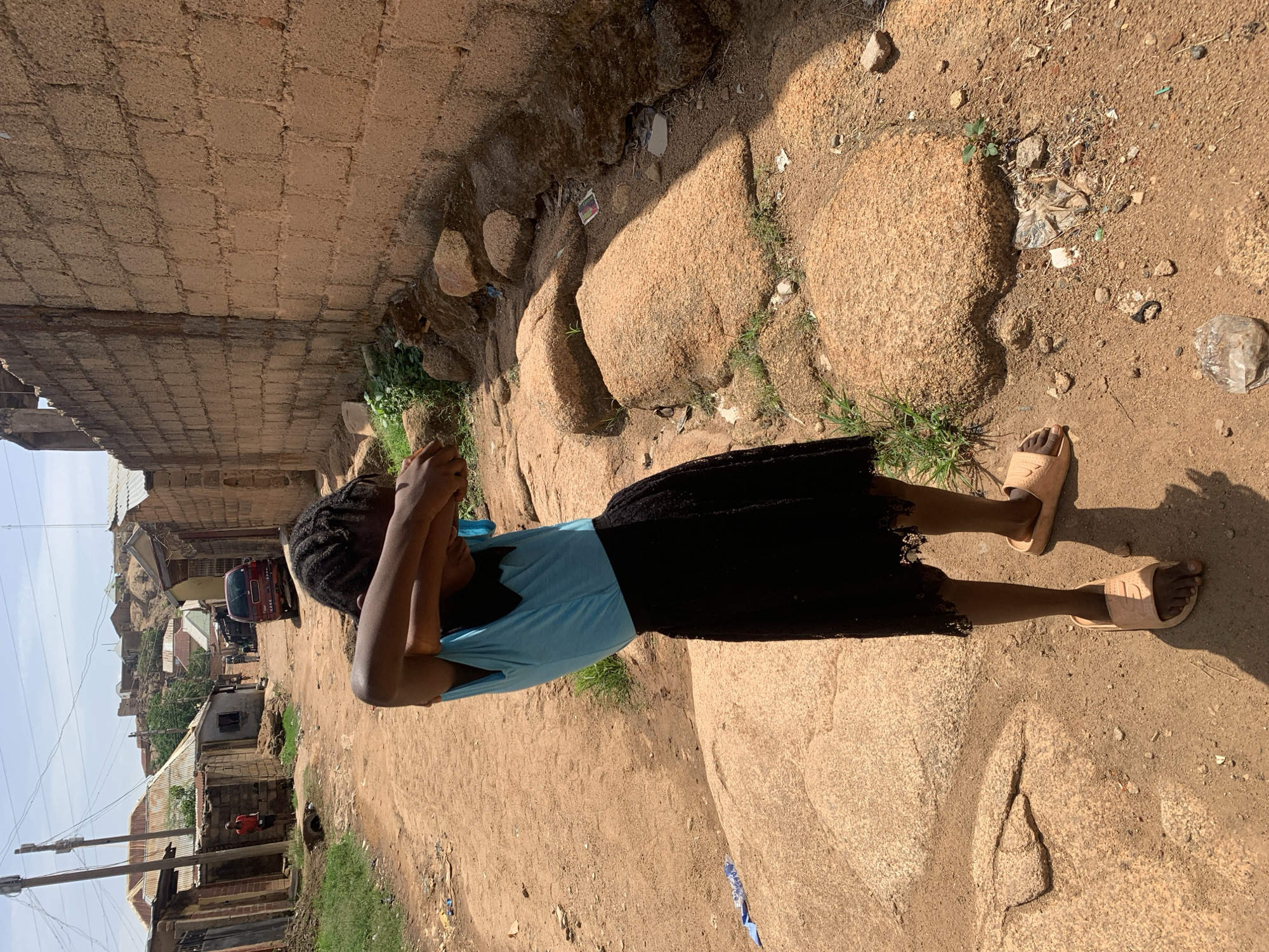 A young Black girl in a light blue shirt and black skirt stands on a rocky, dirt-strewn path, holding her hands to her face. She wears tan sandals. The path is lined with large, smooth rocks and some sparse vegetation. To the left, there's a brick wall, and beyond that, a view of Jos, Nigeria, with buildings clustered together and power lines visible in the sky. A red truck is partially visible on the street in the distance. The overall lighting suggests a sunny day.