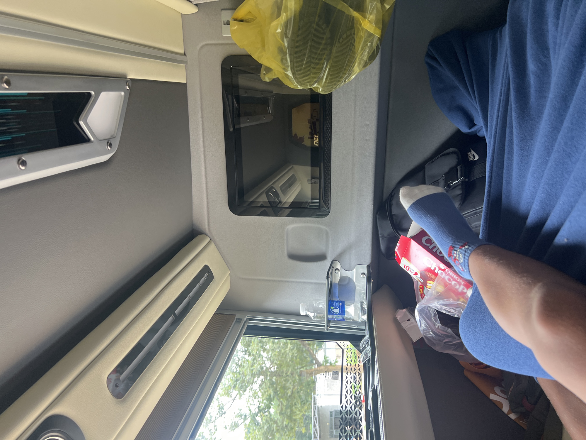 The image shows the interior of a vehicle, likely a bus or van, with a person's legs visible in the foreground, covered by a blue blanket. The person is wearing blue socks with a pattern. Adjacent to the legs is a red and white box of "Choco Pie". To the right, a yellow plastic bag filled with dark items is hanging. A small black screen is mounted on the wall of the vehicle, displaying a blurry image. On the left, a window looks out onto trees and some buildings, suggesting a semi-urban or rural setting. The interior of the vehicle features cream-colored upholstery and air vents. Based on the context provided by the prompt, this scene is located in Pờ Y, Vietnam.