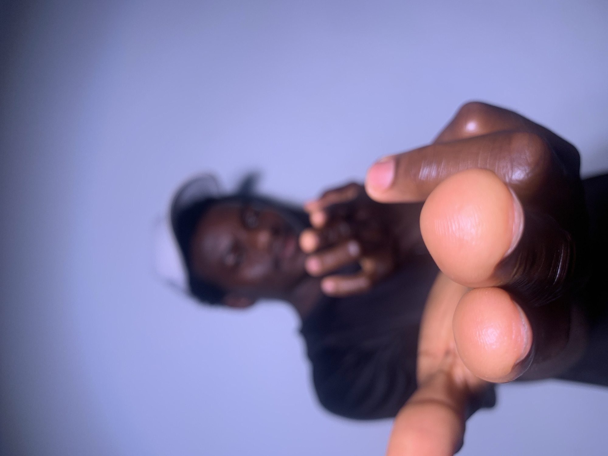 The image features a close-up shot of a person's hands extended towards the camera. The person's face is visible in the background but is out of focus, making it difficult to discern specific features. They appear to be wearing dark clothing. The lighting creates strong highlights on the hands, suggesting a studio setting or controlled lighting conditions. The background is a plain, soft blueish-grey, which further emphasizes the subject's hands. The overall composition is dramatic, with the hands dominating the foreground and creating a sense of direct engagement with the viewer. There is no visible text in the image, and no cues regarding time of day, weather, or specific location are present. The image seems to capture a moment of gesture or interaction, with the outstretched fingers inviting or perhaps reaching out.