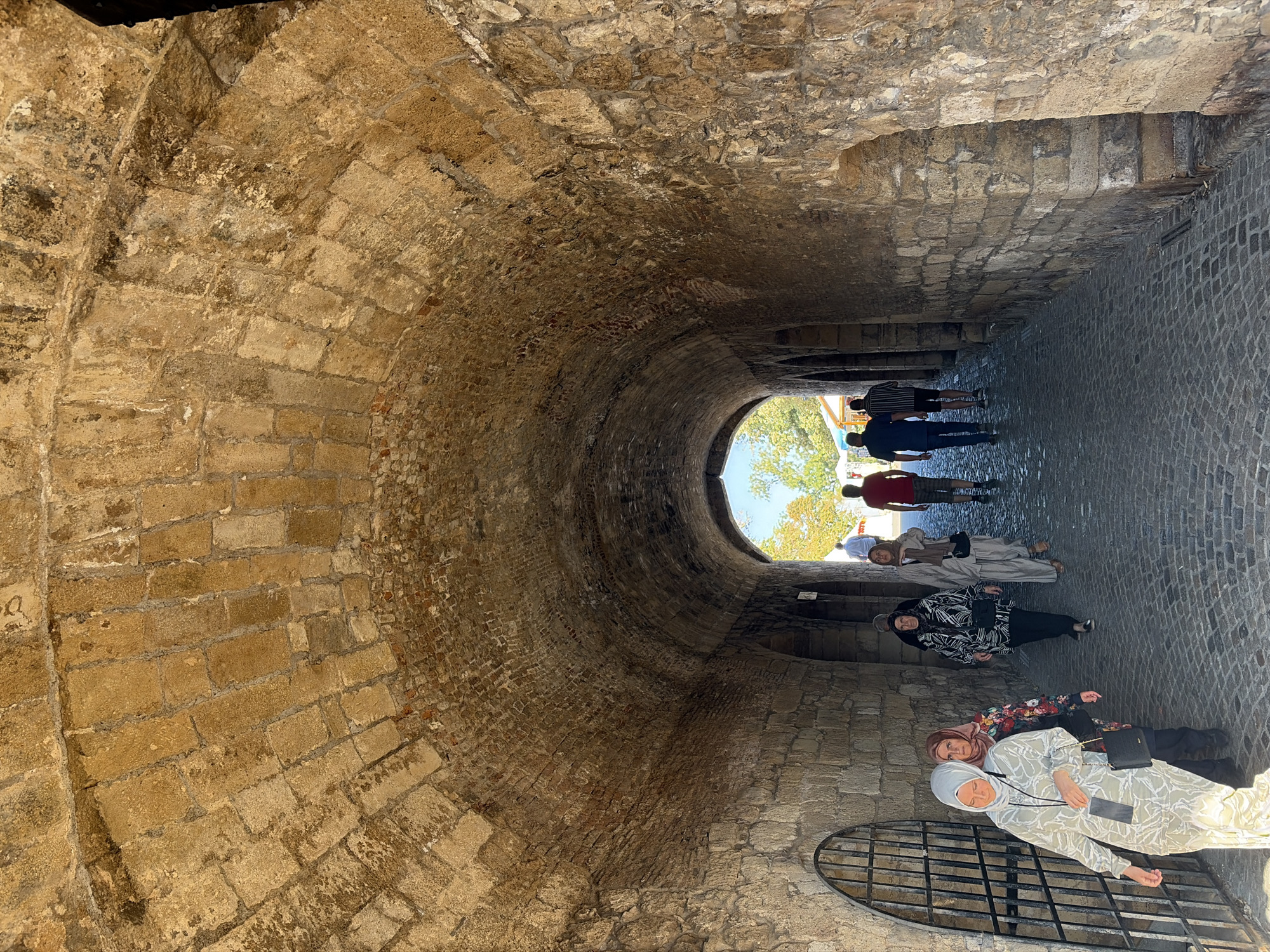The image shows the entrance to the Belgrade Fortress, specifically the access tunnel leading from the Upper Town to the Lower Town in Belgrade, Serbia. The tunnel is constructed of weathered, rough-hewn stone blocks, forming an arched ceiling. Natural light illuminates the end of the tunnel, revealing trees and the sky beyond. Several people are walking through the tunnel, some from left to right, and others from right to left. On the left side of the tunnel, there is a decorative metal gate. The ground is paved with cobblestones.