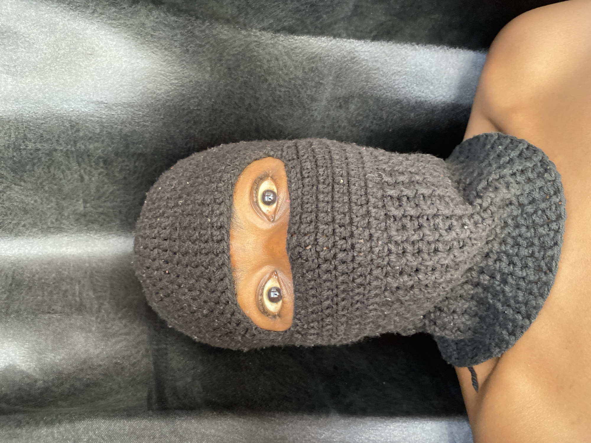 A person wearing a black knitted balaclava is lying down on a black fabric surface. Only the eye area of the person's face is visible through the mask. The person's bare shoulder is visible in the upper right of the image. The image provides no information to determine if this image was taken in Barakin Akawo, Nigeria.