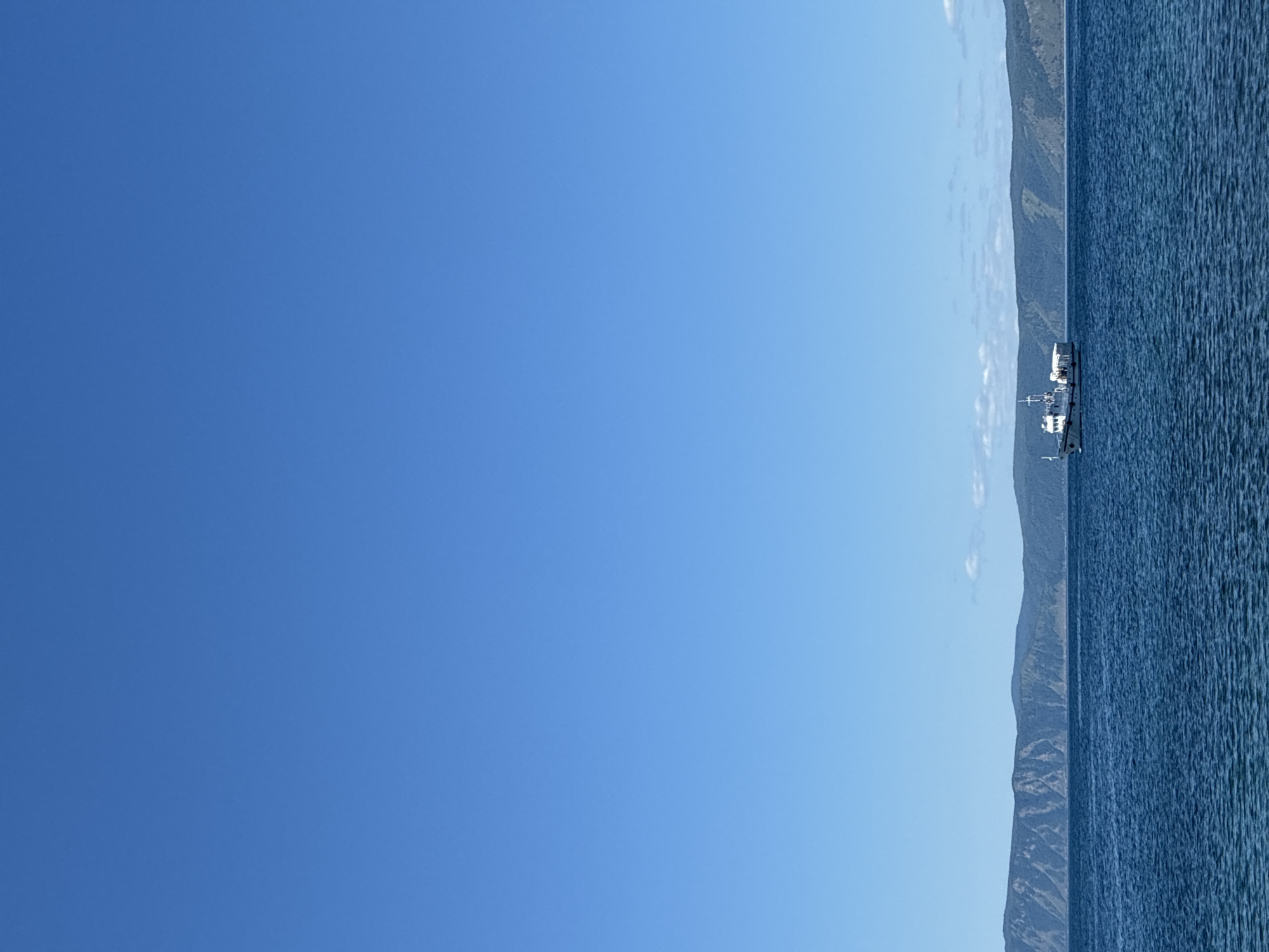 A wide-angle, vertical shot captures a boat sailing on a deep blue body of water. The boat, white with red accents, is positioned slightly to the right of the frame's center and appears to be a passenger vessel, with multiple people visible on its deck. The water exhibits a rippling texture. In the distance, a long, undulating mountain range forms the horizon, its slopes covered in dark green foliage. The sky above is a clear, vibrant blue, devoid of clouds except for a faint wispy trail on the right side, near the mountains. This scene is likely near Listvyanka, Russia, a settlement on the southwestern shore of Lake Baikal, known for its mountainous backdrop and lake excursions.