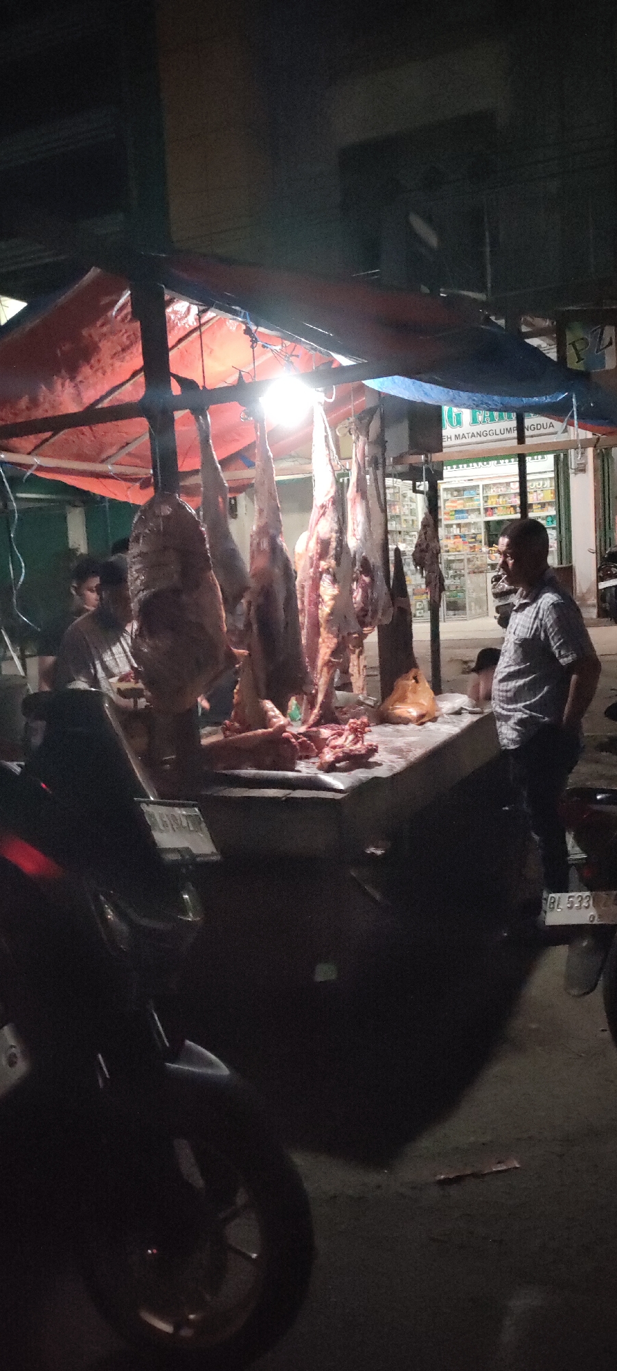 The image captures an outdoor meat stall in Pantegajah, Indonesia, at night. Several animal carcasses are suspended from a makeshift metal frame covered with red and blue tarps. A bright light illuminates the meat and the countertop below. Two men appear to be working at the stall, while another man wearing a plaid shirt stands nearby, possibly a customer. A parked motorbike is visible in the foreground, its license plate partially visible. In the background, across the street, is a pharmacy, identifiable by the sign that reads "EH MATANGGLUMPUNGDUA." The general ambiance is dark, with the stall providing the primary source of light.