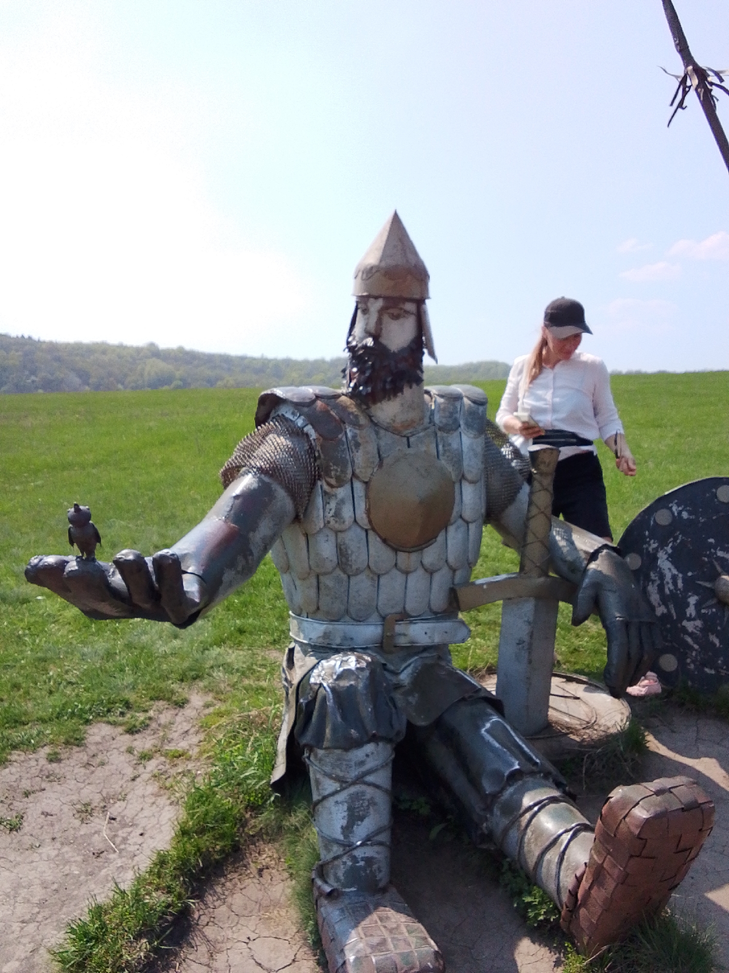 The image shows a metal statue of a medieval knight with a small bird perched on its outstretched hand, located outdoors in a grassy area. The statue is in a seated position, wearing armor and a helmet, and holding a sword and shield. A woman wearing a white shirt and a black baseball cap is standing behind the statue. The background consists of a green field and trees under a blue sky. The location is Каменка, Russia.
