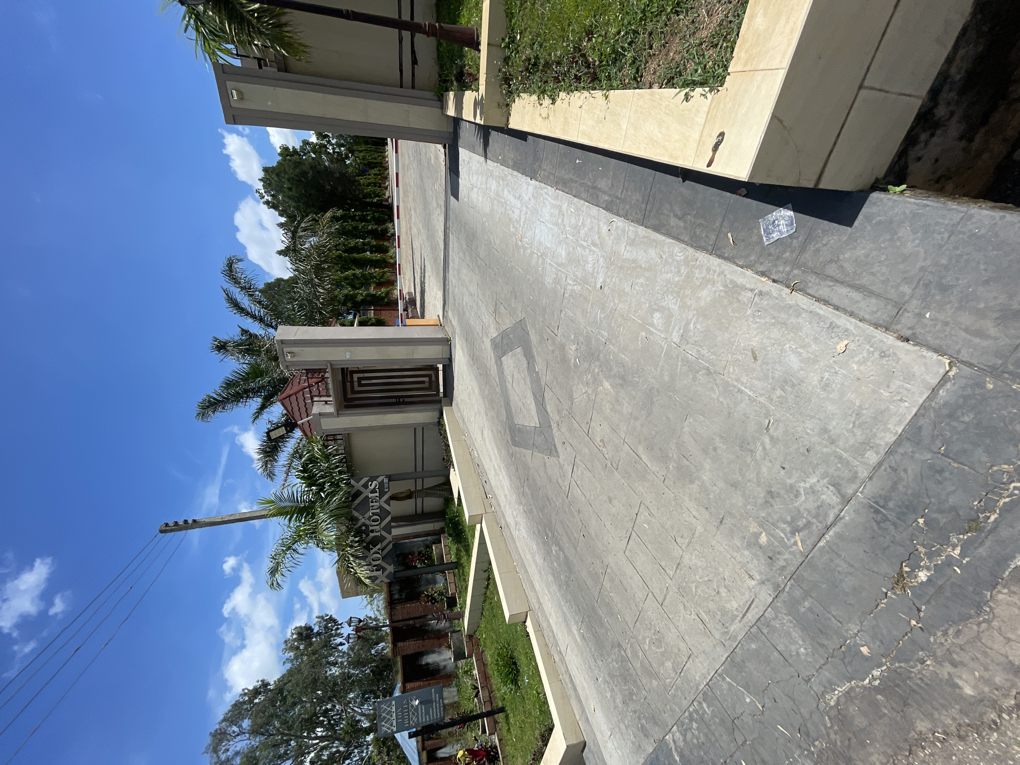 The image captures the entrance to Fox Hotels in Jos, Nigeria, on a bright, sunny day with scattered clouds. The foreground features a grey, patterned concrete driveway leading towards an open gateway. To the left of the driveway, a well-maintained lawn area is bordered by a low, cream-colored wall. Further to the left, a sign displays "FOX HOTELS" and lists services such as "Restaurant," "Bar," "Laundry," "Conference Hall," and "Executive Cream." Palm trees and other lush greenery are visible around the entrance.
The gateway itself is flanked by tall, cream-colored pillars. Through the gateway, a road extends into the distance, lined with trees on both sides. A red and white barrier is visible further down the road, indicating a controlled entry point. The overall impression is one of a clean and organized hotel entrance, set against a vibrant natural backdrop.