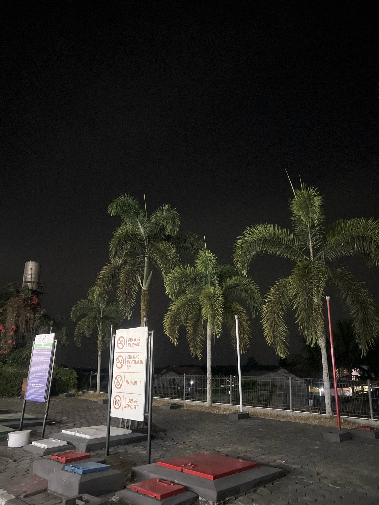 The image shows a nighttime scene in Magelang, Indonesia, likely at a gas station or similar service area. Several palm trees are prominent in the foreground, their fronds illuminated by artificial light. Two information boards are visible, one purple and one white with red lettering. The white board displays four prohibition symbols and text in Indonesian: "DILARANG MEROKOK" (No smoking), "DILARANG MENYALAKAN API" (No open flames), "MATIKAN HP" (Turn off mobile phones), and "DILARANG MEMOTRET" (No photography). The purple board, partially obscured, also appears to contain informational text. In the background, a metal water tower stands near some foliage, and a metal fence runs along the perimeter of the area. Several rectangular access panels with red and blue lids are set into the paved ground. The sky is dark and overcast.
