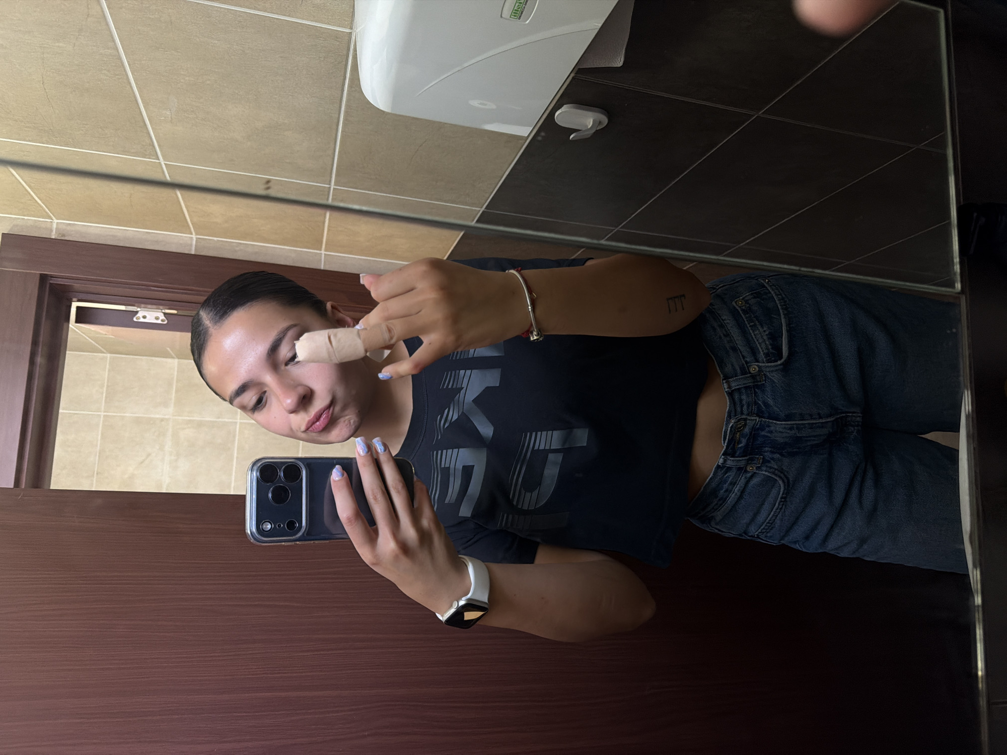 A person is taking a selfie in a bathroom mirror. The person, presumed to be female, is wearing a dark t-shirt, blue jeans, and an Apple Watch. The subject has a bandage on their eyebrow. The bathroom has beige and brown tiled walls, with a white hand dryer mounted on the wall. The reflection shows the person holding a smartphone with multiple camera lenses. There is no visible evidence to determine if this image was taken in the city of Gabrovo, Bulgaria.