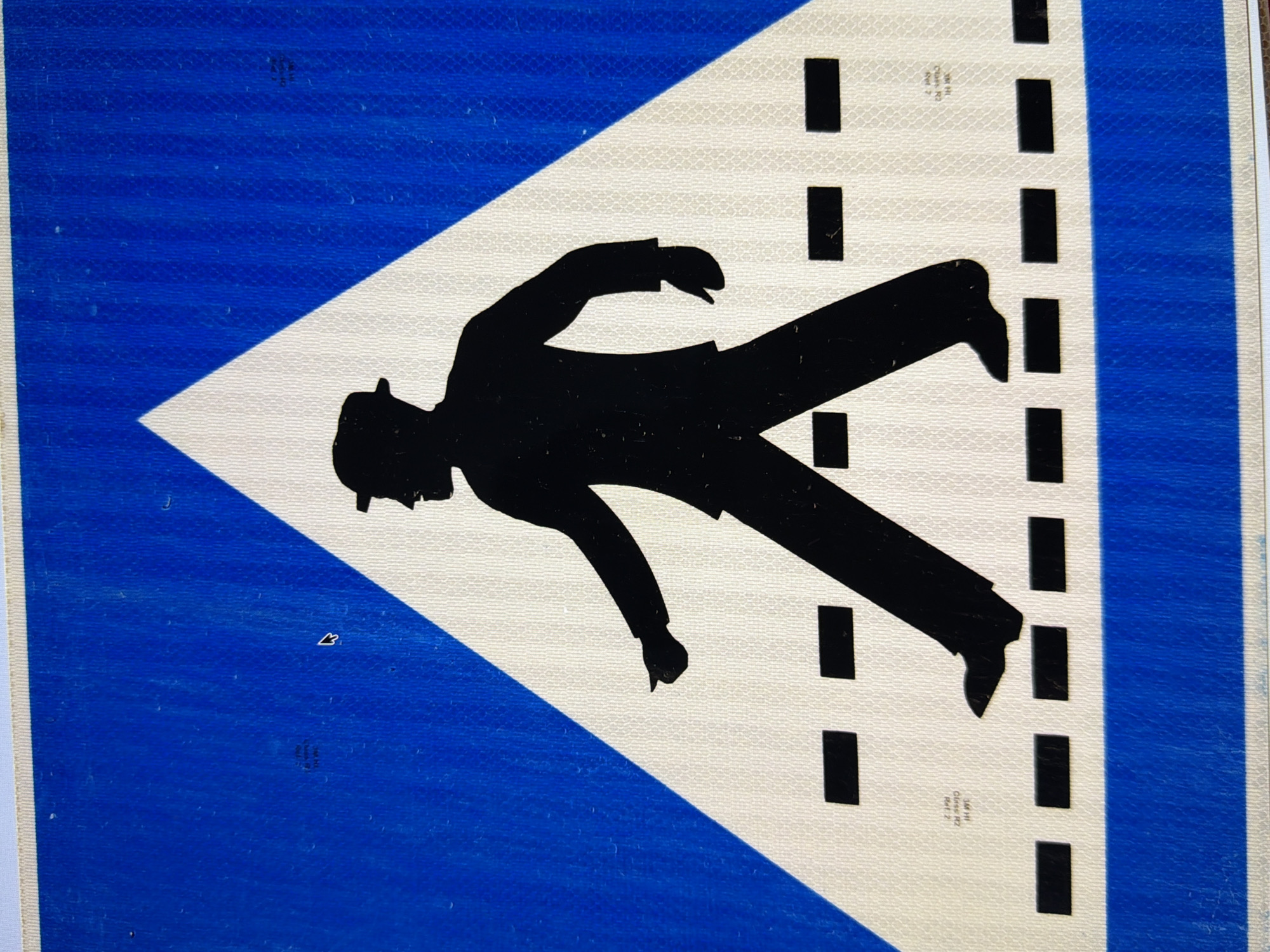 The image shows a close-up of a pedestrian crossing sign. The sign features a black silhouette of a person wearing a hat and suit, walking across a white triangle with a blue background. The pedestrian is depicted mid-stride, with one leg forward and the other back. The white triangle contains a pattern of black horizontal dashes, representing the pedestrian crossing. The bottom portion of the sign is a solid blue band. There are some faint markings on the blue background which appear to be part of the sign's manufacturing or certification information, but they are not clearly legible. The image is taken at a slight angle, suggesting it is a photograph of a physical sign. Without further context or information, the specific location in Kyiv, Ukraine, cannot be determined from the image alone.
