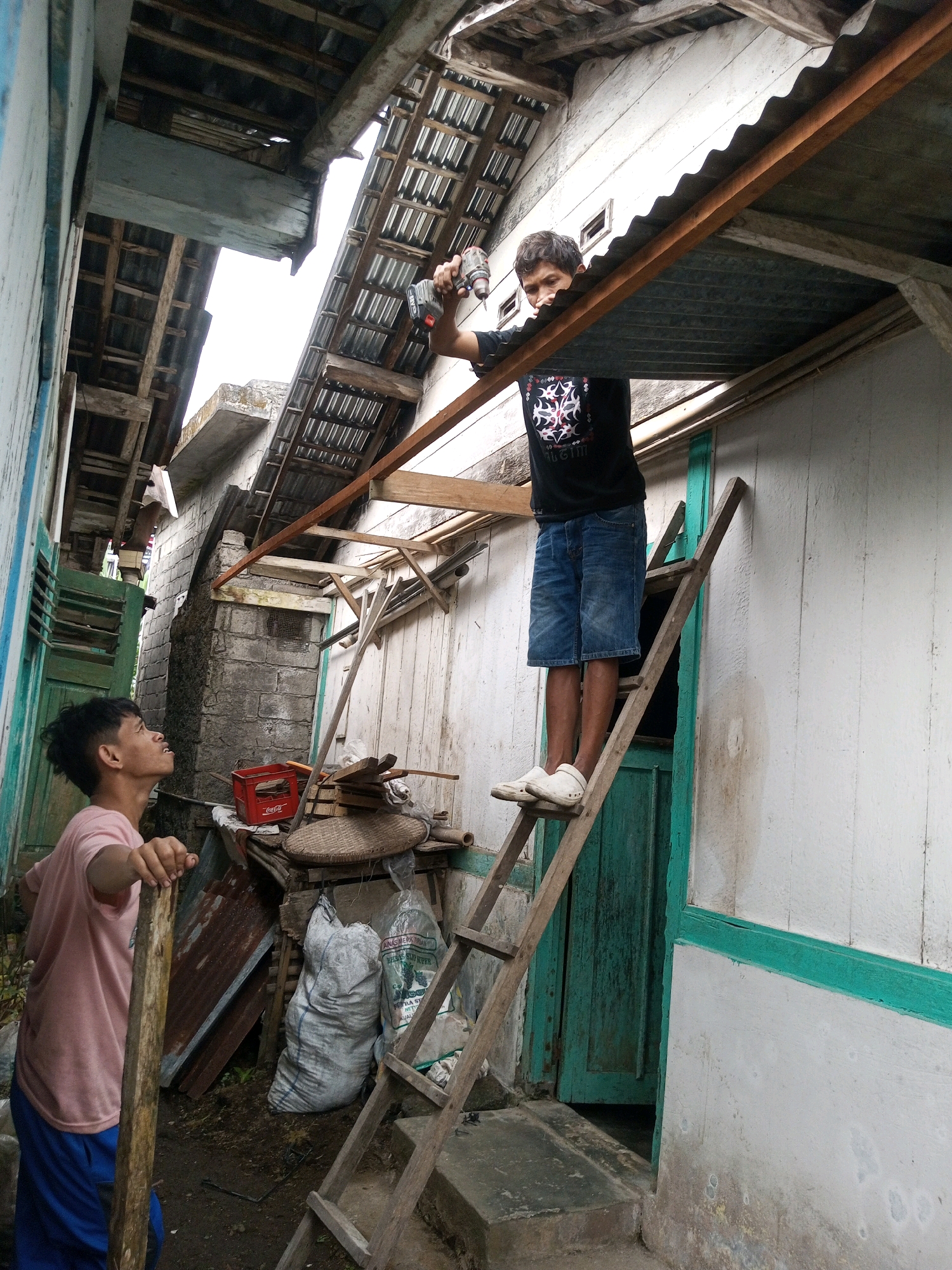 In Andongsili, Indonesia, two men are working on the roof of a building. The man on the ladder, wearing a black shirt and blue shorts, is using a cordless drill to attach a metal sheet to the roof. The other man, wearing a pink shirt and blue pants, is standing below and holding a wooden pole, looking up at the man on the ladder. The scene appears to be in a cluttered outdoor area with various construction materials and household items visible.