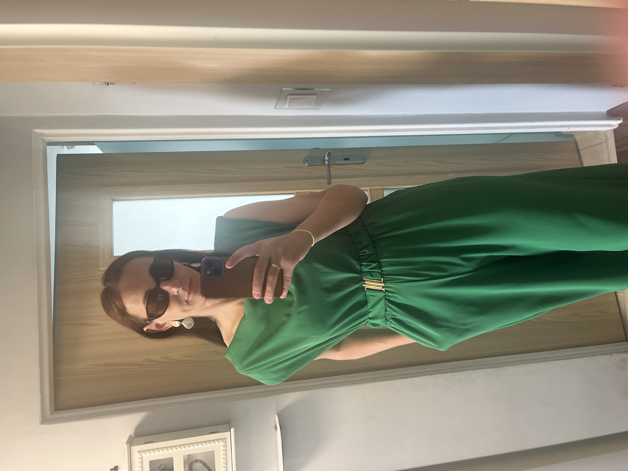 A woman stands in a hallway, taking a selfie in a mirror. She is wearing a green jumpsuit with a gold belt and large, teardrop-shaped earrings. She also wears dark sunglasses. The hallway has a light-colored door and a white wall with a light switch.