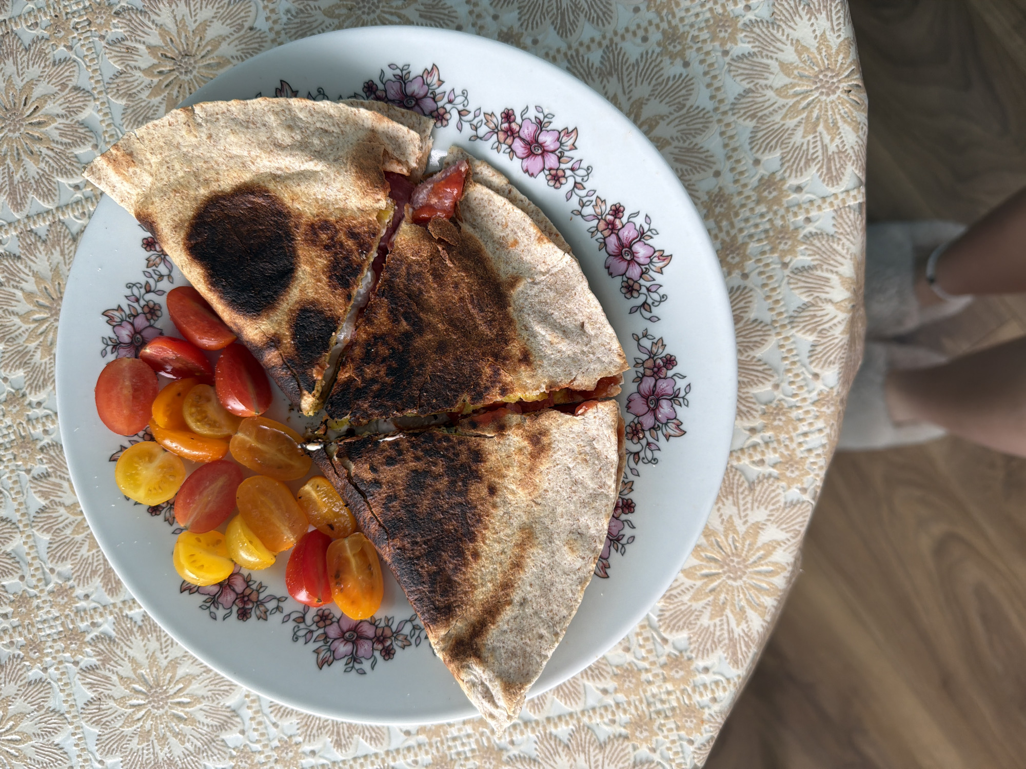 A top-down shot displays a white plate with a floral design on a tan, textured tablecloth. The plate holds a pita-like bread, sliced into two triangular pieces, with dark spots, seemingly from grilling. Beside the bread are halved cherry tomatoes of red, orange, and yellow colors. A person wearing white slippers stands adjacent to the table with the plate. The location is unknown but is not Varna, Bulgaria, as the image could have been taken anywhere.
