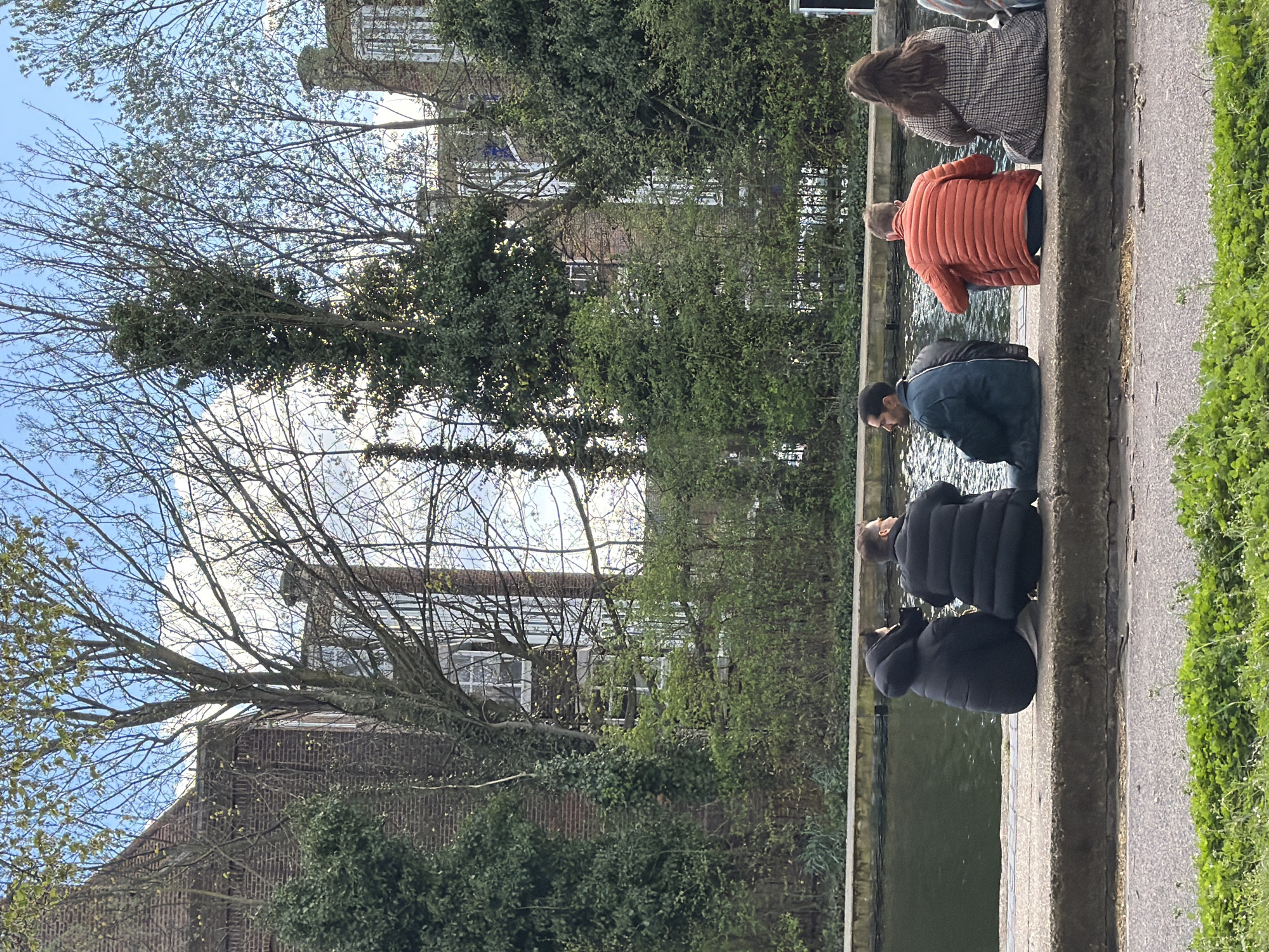 Several people are sitting on the edge of a canal or waterway. Four figures sit on the edge, wearing coats suitable for cool weather. The person closest to the viewer wears a black, puffy coat. Next to them is a person in a dark blue jacket, an orange jacket, and a brown checkered coat. They are looking into the water. Lush greenery and trees line the canal. In the background are brick buildings with glass windows and roofing. The sky is partly cloudy. This appears to be a scene within the city of London, United Kingdom.
