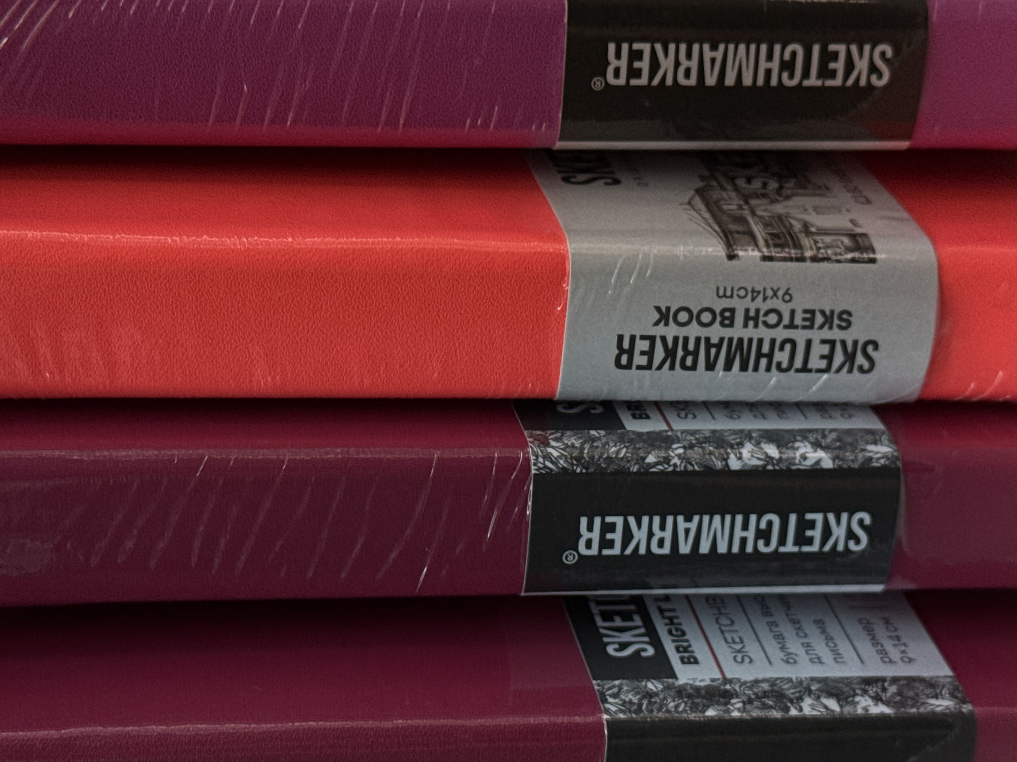 The image displays a close-up view of four sketchbooks, side by side, with their spines facing the camera. The sketchbooks are wrapped in transparent plastic. The first sketchbook from the left is a deep pinkish-purple, followed by a vibrant red one, and then another deep pinkish-purple one. The fourth sketchbook, partially visible on the right, appears to be a darker shade of purple. Each sketchbook has a label wrapped around its lower section. The label on the first sketchbook has "SKET" visible, and below it, "BRIGHT L", "SKETCHB", "бумага выс", "для скетч", "письма", and "размер 9x14 cm". The label on the second sketchbook reads "SKETCHMARKER", "SKETCHBOOK", and "9x14cm". The label on the third sketchbook also has "SKETCHMARKER" written vertically. The background is blurred, suggesting a retail environment, possibly a store shelf. There are no people or specific city landmarks visible in the image. The lighting is even, highlighting the textures and colors of the sketchbooks.