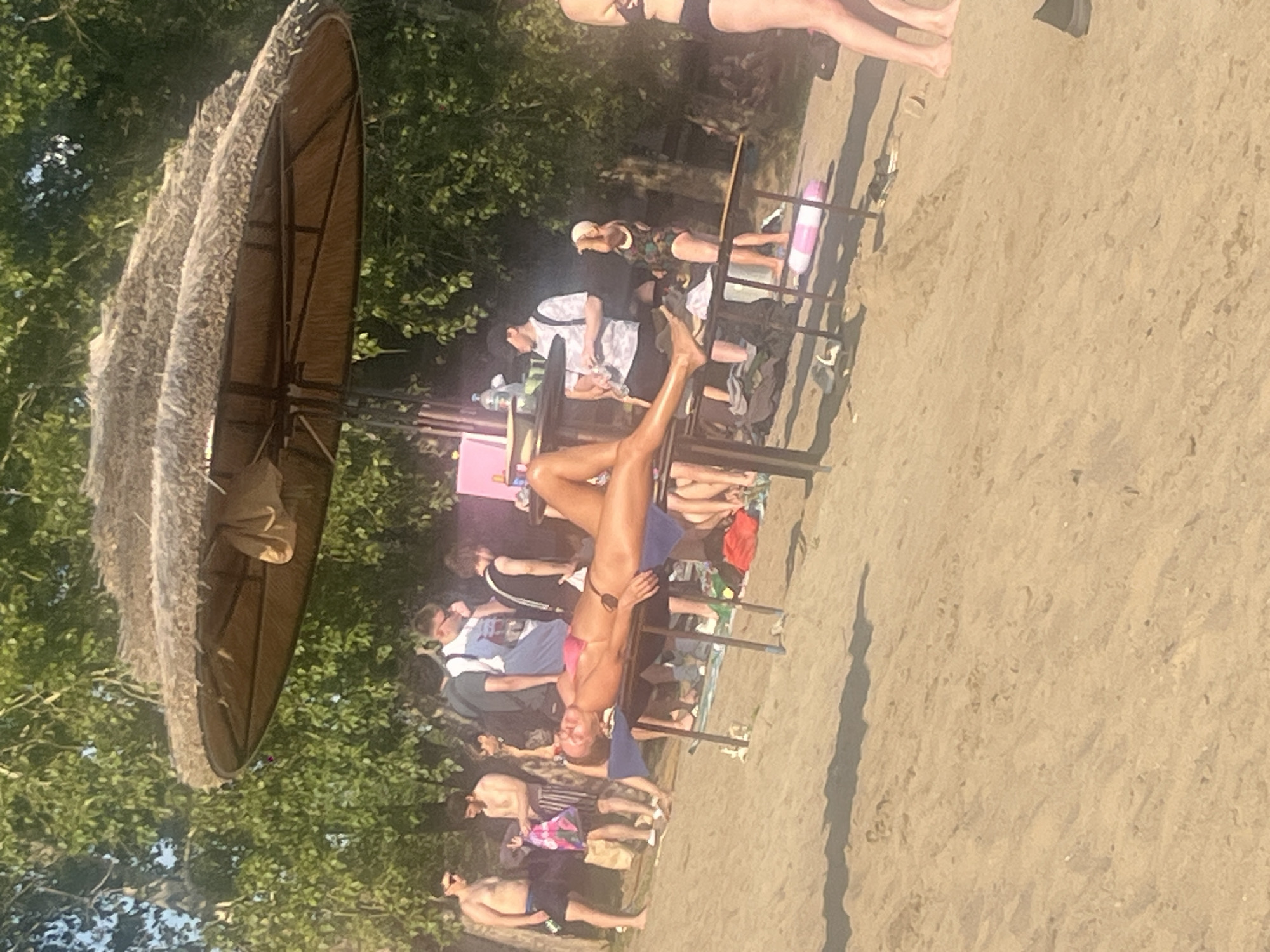 A woman in a pink bikini is sunbathing on a table at a beach. She is lying on her back with her head slightly tilted to the side. There are other people in the background, some standing and some sitting. The scene appears to be a public beach with trees and a thatched umbrella providing shade. The ground is sandy. The image does not provide any specific details or context that would allow for identification of the location within the city of Tyumen, Russia.