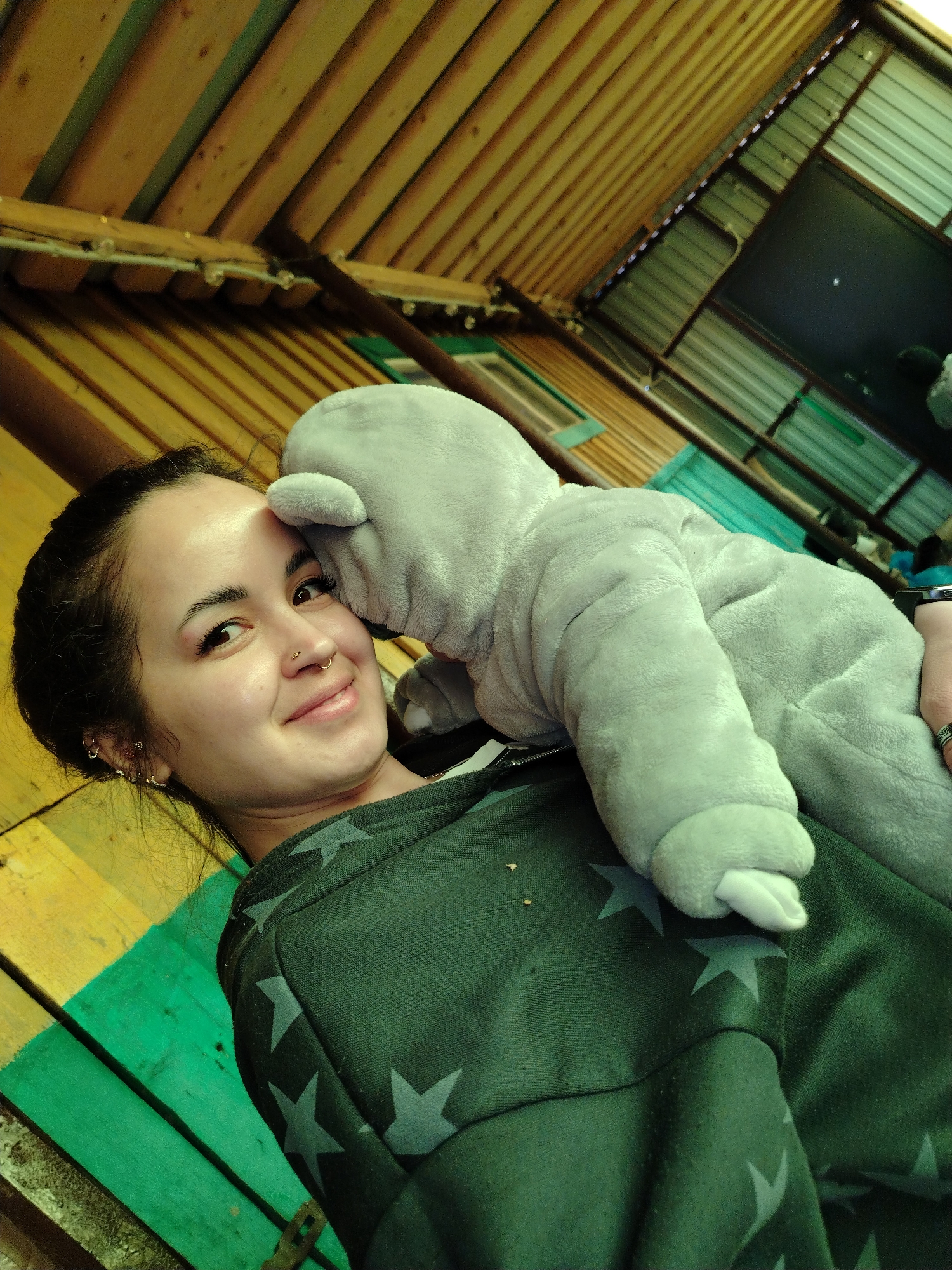 A young woman with dark hair pulled back from her face smiles at the camera while a plush gray hippo is pressed against her cheek. She has a nose ring and multiple earrings. She is wearing a dark green jacket with gray stars. The scene is outdoors, underneath a wooden and metal structure, likely a patio or porch. The wooden beams above are light brown, and the metal panels are green and silver. The location is Гожан, Russia.
