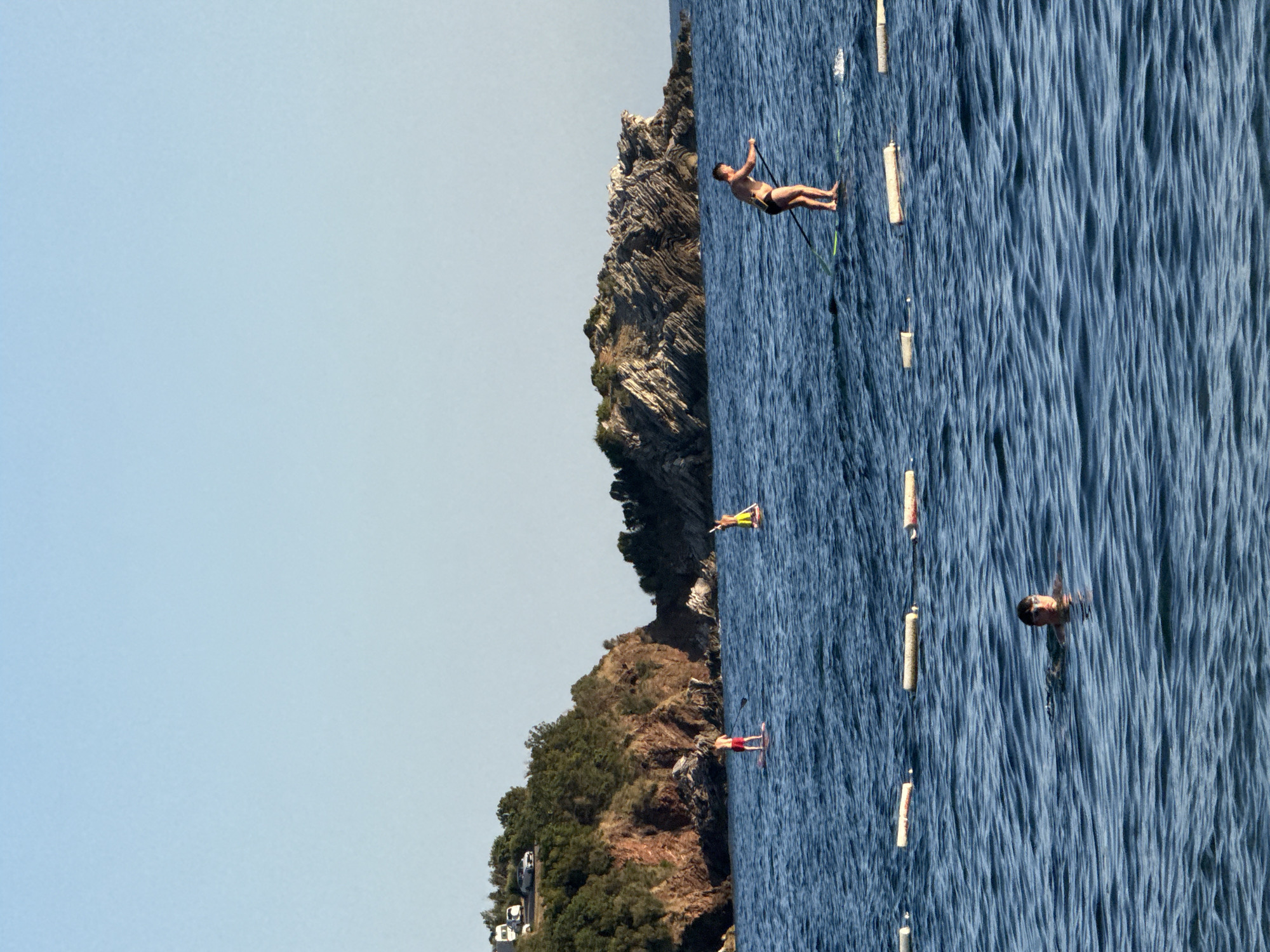 In Pržno, Montenegro, on a clear, sunny day, a scenic view of the Adriatic Sea unfolds. The calm, blue waters are dotted with several individuals enjoying water activities. In the foreground, a person swims with their head above water, wearing goggles. Further out, two men are stand-up paddleboarding. One is closer to the shore, standing on a light-colored paddleboard, while the other is further out on a green paddleboard, wearing dark swim trunks. A line of white buoys marks a boundary in the water, separating different areas. In the background, a rocky, tree-covered peninsula juts out into the sea, with a few cars and a camper visible on a road near its summit. The sky is a clear, pale blue, providing a serene backdrop to the coastal landscape.