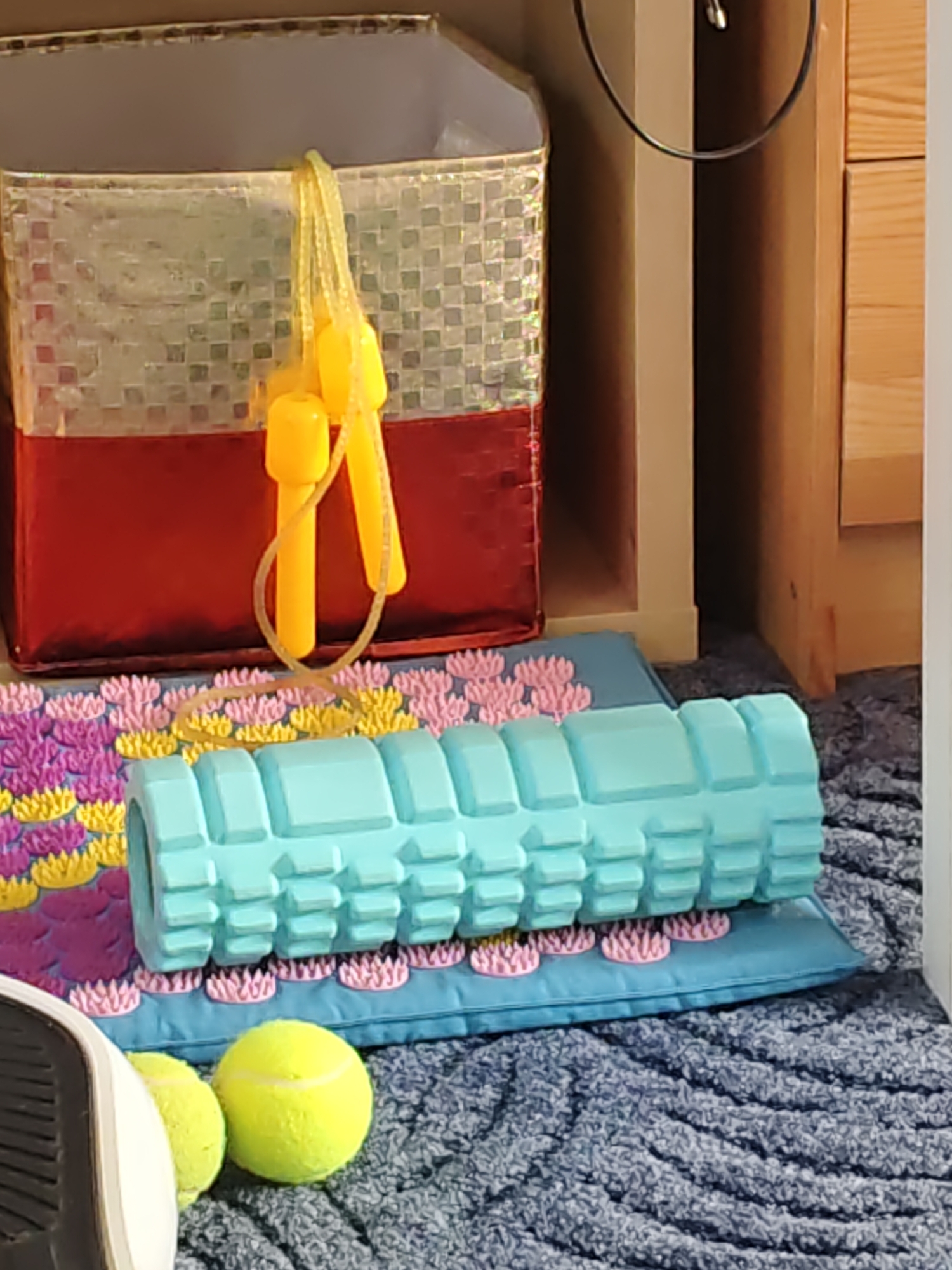 The image displays a collection of fitness and relaxation items arranged on a dark blue, textured surface, possibly a rug. In the foreground, two bright yellow tennis balls are visible. To the left, a portion of a white and black fitness device, resembling an elliptical or treadmill foot pedal, is in the frame. Centered in the mid-ground is a light blue foam roller with a textured surface, resting on a blue mat. This mat is placed on top of a pink and yellow acupressure mat, which has a floral pattern. In the background, a red and metallic silver storage bin with yellow handles from a jump rope is positioned. The scene appears to be indoors, suggesting a home or gym environment. There is no discernible information within the image that would allow for the determination of its location in Rostov-on-Don, Russia.