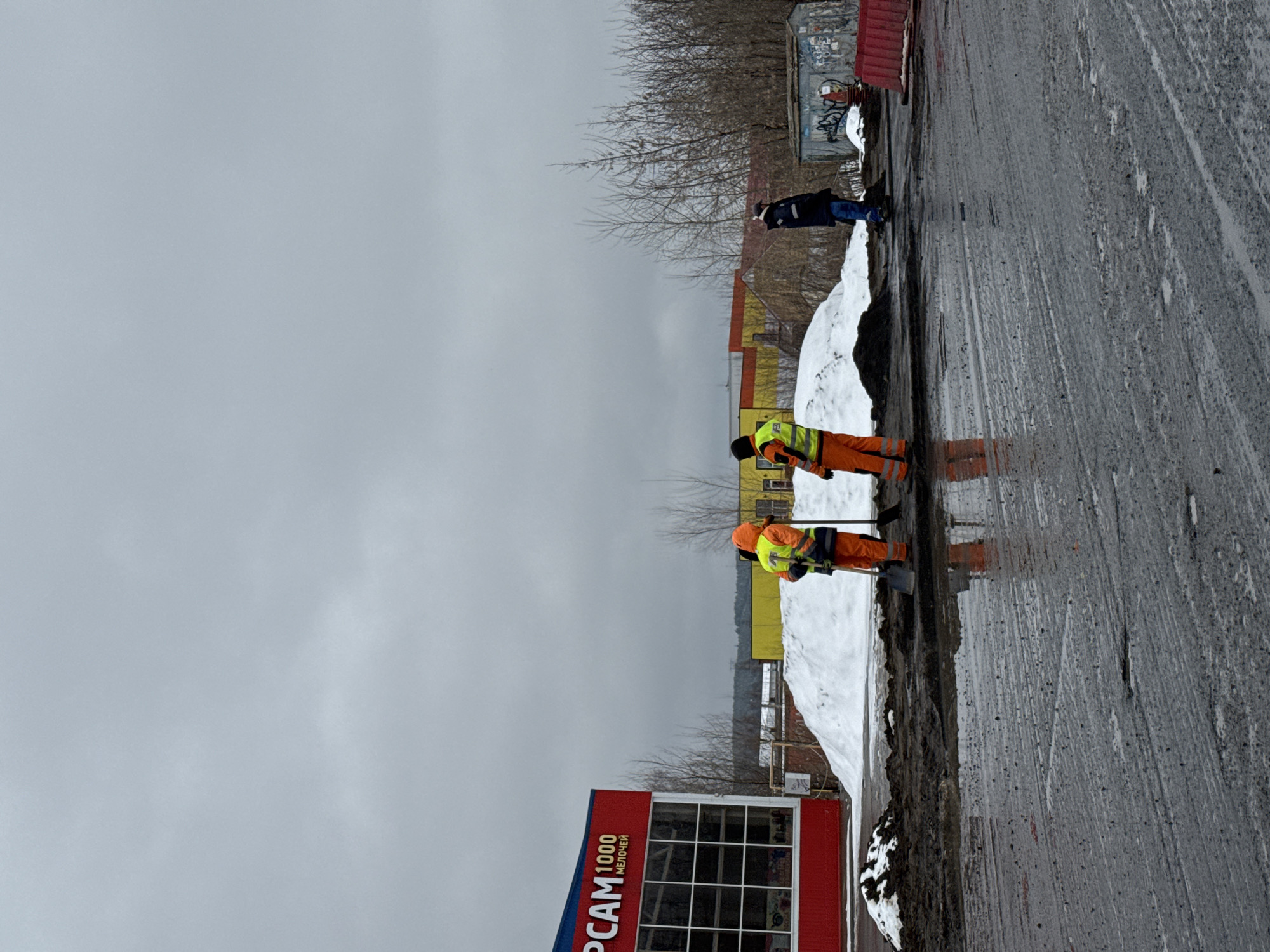 The image shows a street scene in Chaikovsky, Russia. Two workers in high-visibility orange overalls and vests are shoveling snow near the edge of a road. A third person is further away, also on the side of the road, dressed in blue. A building with a red facade and the words "PCAM 1000" in Cyrillic script is visible on the left side of the image. Snow piles line the roadside. The sky is overcast, and the road surface is wet. In the background, there are trees without leaves, buildings of varying colors, and a structure with graffiti. The road surface shows signs of wear and tear. The location appears to be an urban area, possibly undergoing snow removal efforts.
