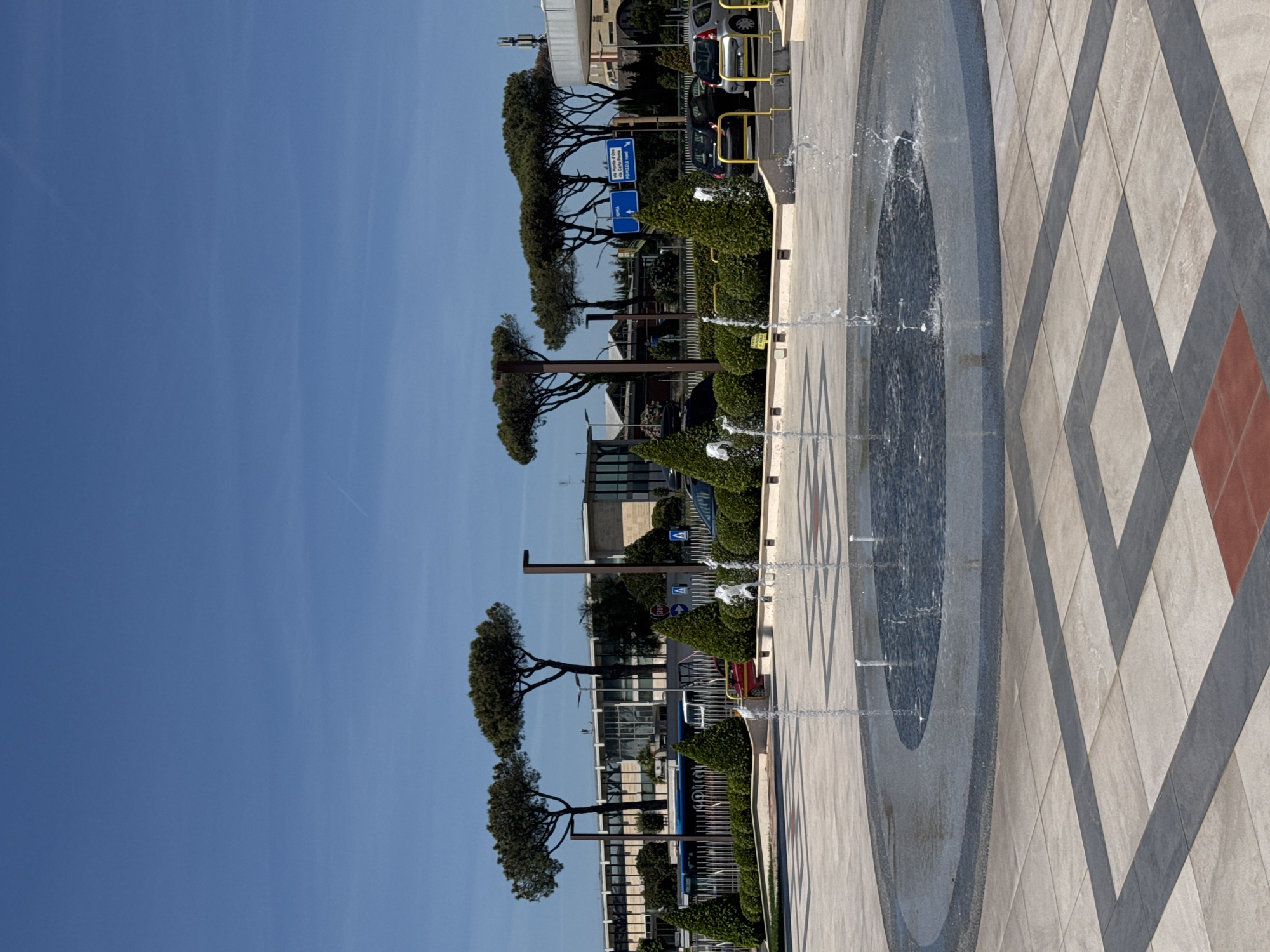 The image depicts a modern urban space in Pomezia, Italy. In the foreground is a circular water feature with jets of water spraying upwards. The surrounding pavement is designed with a geometric pattern of alternating light and dark gray tiles, accented by a reddish-brown square. Along the edge of the water feature, there is a row of neatly trimmed green hedges. Beyond the hedges, there are slender trees lining the street, with street signs visible. Buildings of modern architectural style can be seen in the background. Several cars are parked near the buildings. The sky is clear and blue.