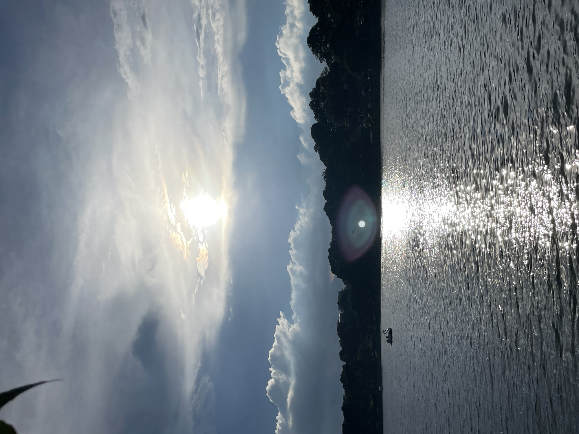 The image depicts a serene lake scene with a treeline of dark green foliage and scattered buildings along the distant shore. The sky is partially clouded, with the sun shining brightly through a gap in the clouds, casting a strong reflection on the rippling water. The foreground is dominated by the lake's surface, showing subtle waves and sparkling sunlight. On the left side of the lake, a swan-shaped paddle boat with several occupants is visible. The overall atmosphere is tranquil, suggesting a late afternoon or early evening setting. This scene is characteristic of Xuân Hương Lake in Đà Lạt, Vietnam, a popular tourist destination known for its natural beauty and recreational activities.