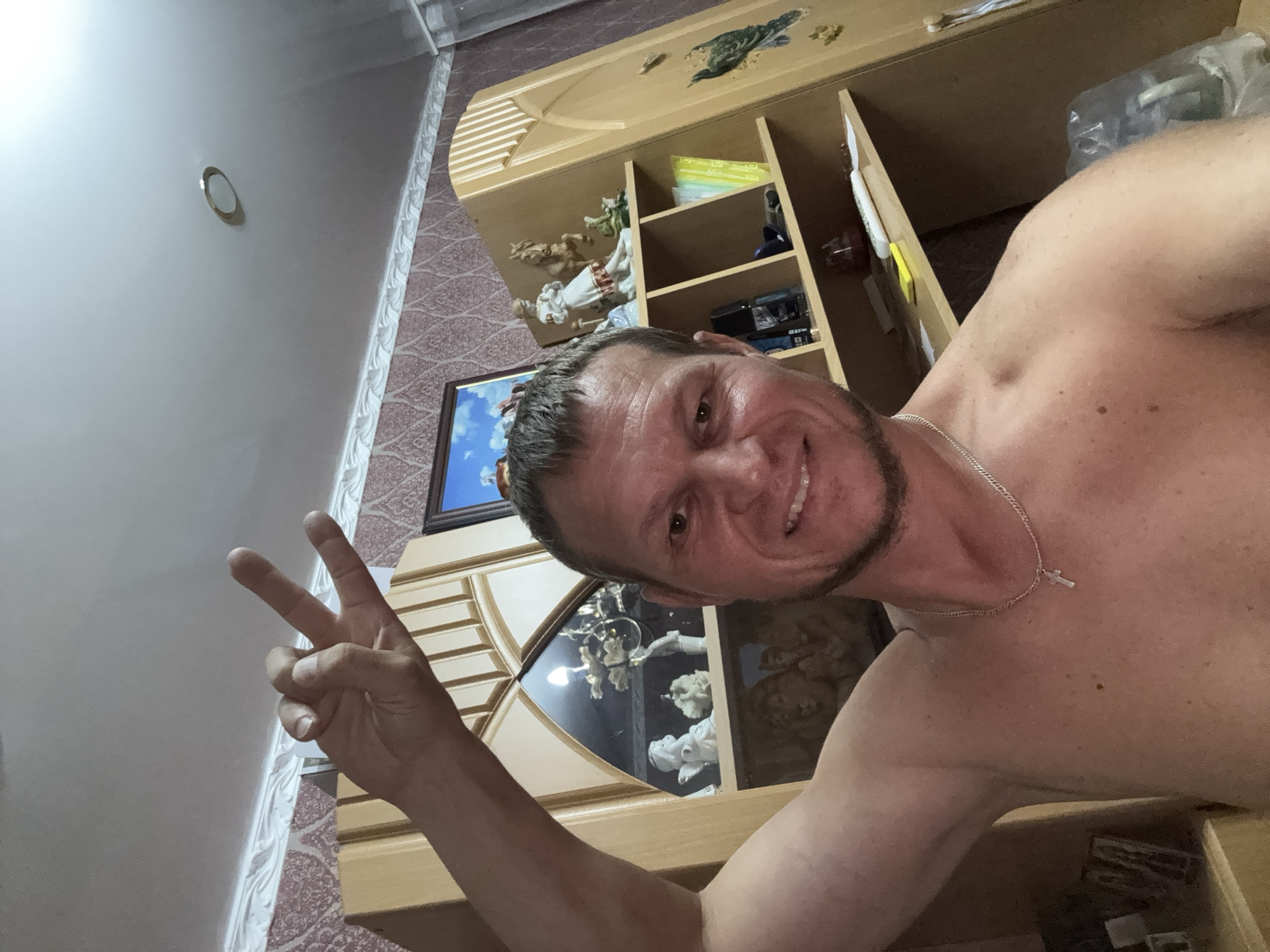 The image shows a shirtless man with a short beard, wearing a silver chain with a cross pendant, taking a selfie in what appears to be a home interior. He is smiling and holding up his right hand, with his index and middle fingers extended in a peace sign. In the background, there is a wooden cabinet with shelves displaying various decorative items, including figurines and what looks like a framed picture. The room has patterned wallpaper and a white ceiling with a recessed light fixture. There is no visible indication of the city of Glubokoe, Kazakhstan, in the image.