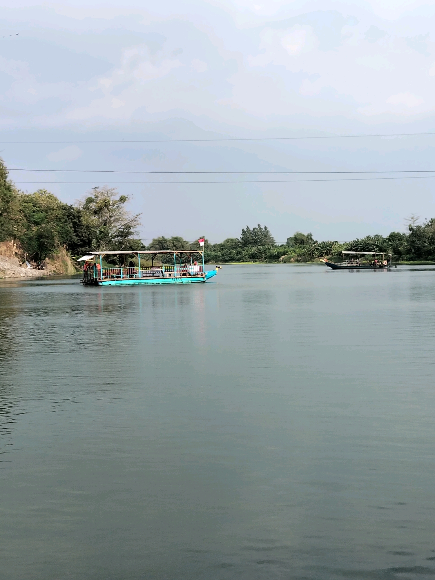 Two boats are visible on a wide, calm river. The boat in the foreground is turquoise with a canopy and appears to be carrying passengers. It is moving from left to right across the frame. A national flag, red and white, is mounted on the rear of this boat. The second boat, further in the distance on the right, is dark-colored and also appears to have passengers. The riverbanks are lined with trees and vegetation, with some rocky terrain visible on the left bank. The sky is overcast. The scene is likely located in or around the city of Kesamben, Indonesia, given the presence of typical Indonesian flora and the style of the boats.