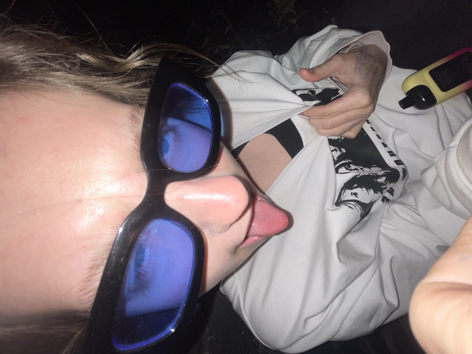 A person is depicted in a close-up shot, angled sideways, in what appears to be a low-light indoor setting. The individual's head is turned, showing a profile view. They are wearing large, cat-eye style black sunglasses with intensely reflective blue lenses, through which a blue eye and eyelashes are visible. The person has light brown or blonde hair, slightly wavy, framing their face. Their tongue is extended slightly from their mouth in a playful manner.
The subject is dressed in a loose-fitting white long-sleeved garment, possibly a t-shirt or sweatshirt, which features a black graphic print on the chest, appearing to be a stylized face or figure. One hand is seen pulling at the white garment, revealing a black undergarment beneath. The person's other hand is visible further up, resting on the white sleeve, and features faint, handwritten text or markings on the back of the hand and wrist. A yellow and pink vape device is casually placed on the white fabric in the upper right portion of the image. The background is dark and out of focus, providing no specific environmental details. The overall impression is casual and somewhat informal. No specific location cues are visible in the image itself, apart from the provided context of Moscow, Russia.