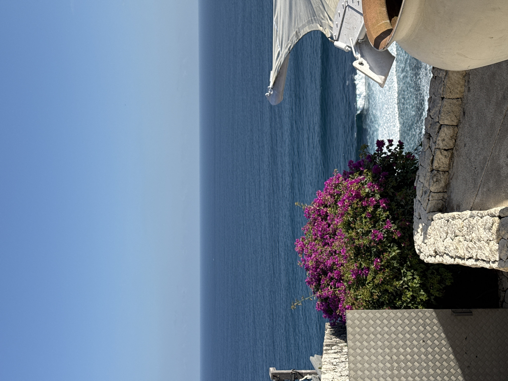 The image captures a scenic view overlooking the ocean, likely from an elevated position in Kuta Selatan, Indonesia. The sky is a clear, vibrant blue. The ocean below is a deep, tranquil blue, with gentle waves creating white foam along the shore. In the foreground, a dense bush with bright purple bougainvillea flowers adds a splash of color. To the left, a textured metallic surface, possibly a door or panel, is visible. The foreground is bordered by stone walls. On the right side, a large, light-colored ceramic pot is partially visible, with a hint of a terracotta-colored object inside. A white awning structure extends from the right, curving towards the ocean. The overall impression is one of a peaceful, coastal setting.
