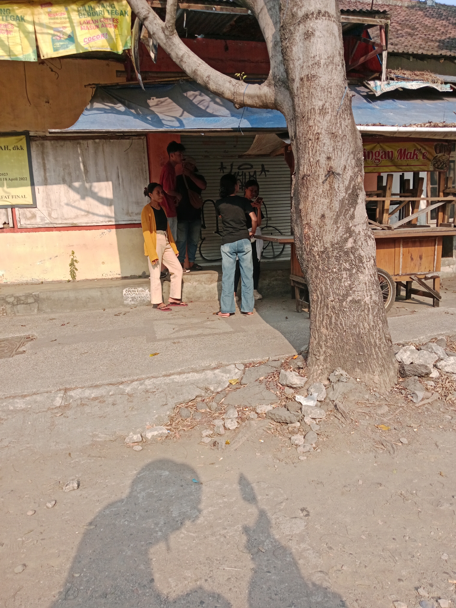 The image captures a street scene in Kediri City, Indonesia, with a focus on a large tree trunk in the foreground. Behind the tree, a group of four individuals are gathered on a raised concrete area, which appears to be a sidewalk or step. Three of the individuals are standing, while one is seated. The individuals are dressed in casual clothing.
To the left of the group, a yellow banner with text is partially visible. Behind them, a building with a corrugated metal roof and shuttered windows forms the backdrop. Some of the shutters are closed, while others are partially open. A wooden cart with wheels is positioned to the right of the group, also against the building.
The ground in the foreground is a mix of concrete and dirt, with scattered rocks and dry leaves. The lighting suggests it is daytime, with shadows cast by the tree and the people. The overall impression is of an everyday street scene in an urban environment.