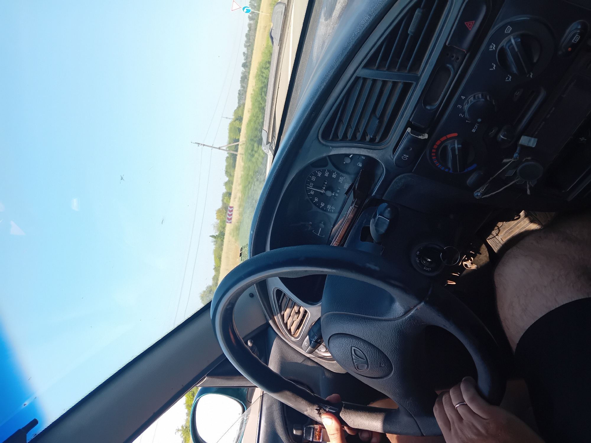 The image shows the interior of a car with a person driving. The steering wheel is in the foreground, and the dashboard with instruments and air vents is visible. The driver's hands are on the steering wheel. The windshield shows a clear blue sky and a rural landscape with fields and trees. The location is identified as Rohan, Ukraine. The scene depicts a moment of driving on a sunny day.