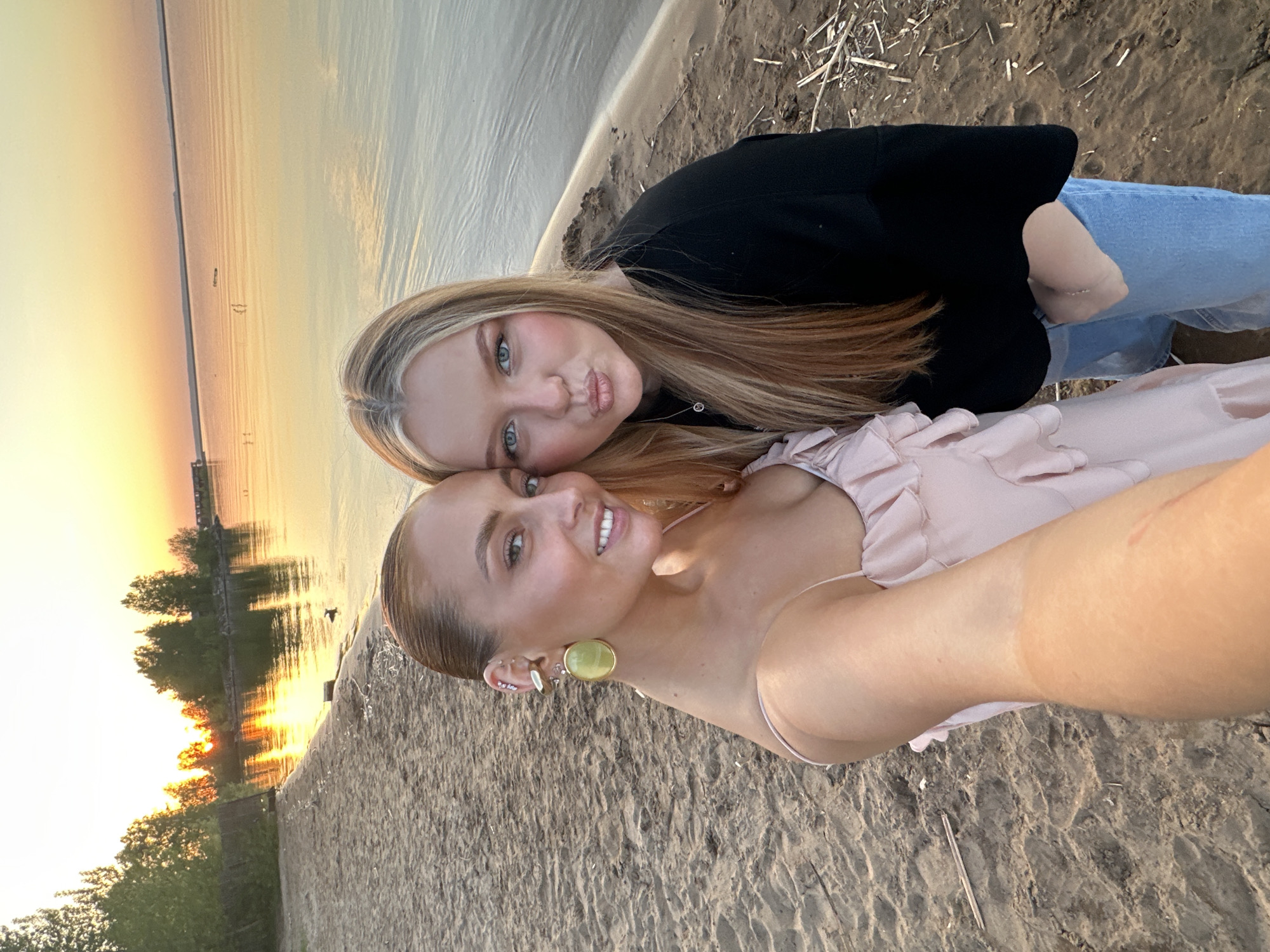The image captures two women posing for a selfie on a beach. The woman in the foreground wears a light pink dress and has multiple earrings. The other woman, positioned behind, wears a black top and light blue jeans. The background shows a body of water reflecting the golden hues of a sunset, with trees visible on the opposite shore. The setting is likely a beach area in the city of Chaikovsky, Russia.