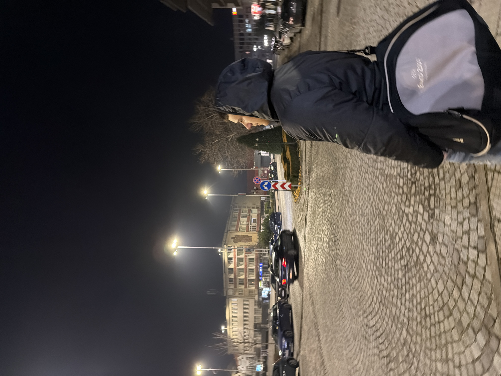 The image captures a person wearing a hooded jacket and carrying a backpack, walking on a cobblestone street at night. The person is facing away from the camera, their profile visible under the hood. The street appears to be in an urban setting with multiple street lights illuminating the area. Parked cars line the side of the street, and buildings are visible in the background. The image seems to be taken at night, given the dark sky and the artificial lighting. The road signs suggest that this scene is located in the city of Panagyurishte, Bulgaria.