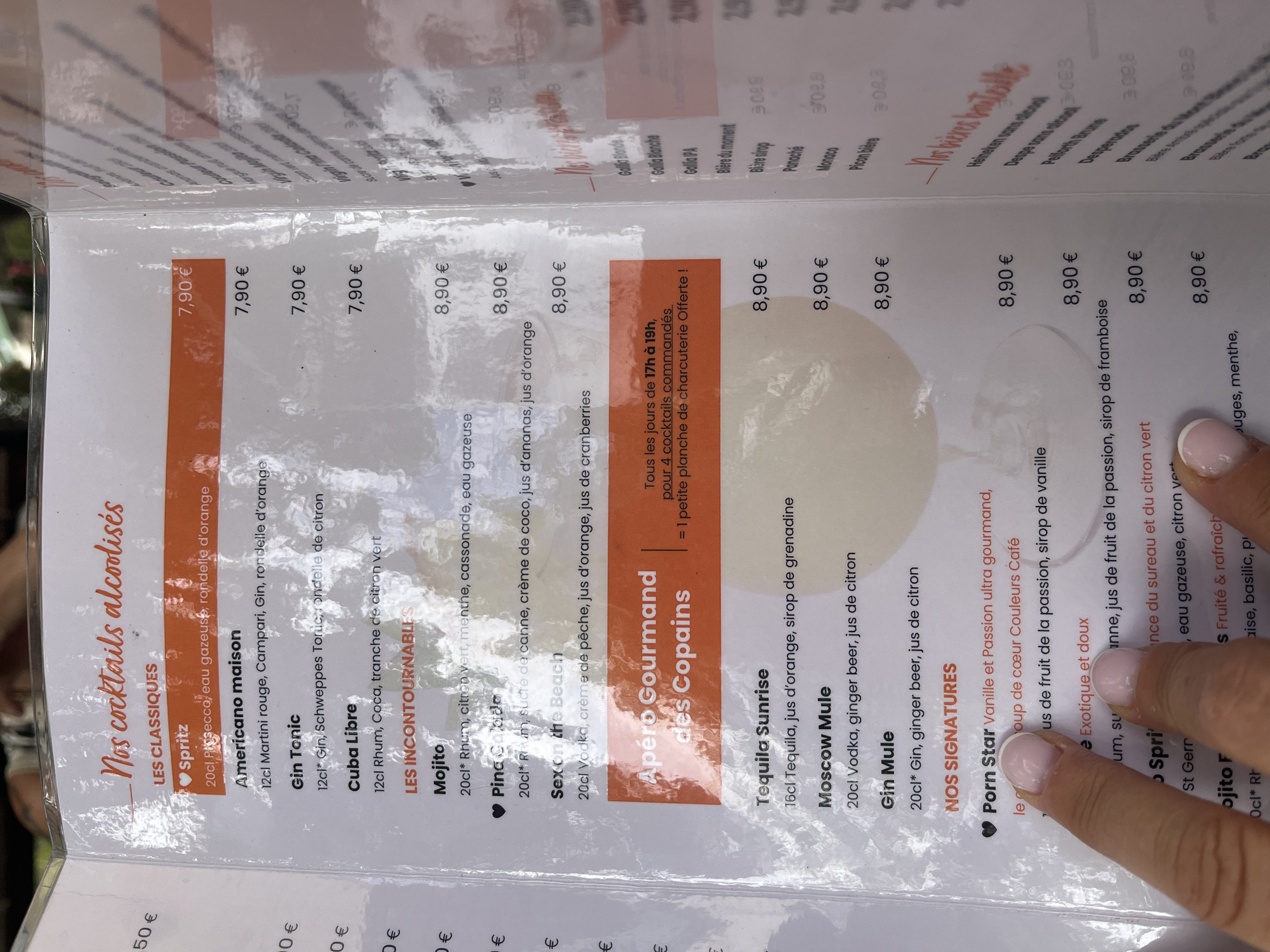 The image shows a close-up of a menu from a restaurant or bar, likely in Montélimar, France, as indicated by the currency symbol "€" and French text. The menu is open, with the left page featuring a list of alcoholic cocktails divided into "LES CLASSIQUES" (The Classics) and "LES INCONTOURNABLES" (The Must-Haves). Prices are listed in Euros. Some of the classic cocktails include Spritz, Americano maison, Gin Tonic, and Cuba Libre. Under the "Must-Haves" section, Mojito, Pina Colada, and Sex on the Beach are visible. There is also a section titled "Apéro Gourmand des Copains" with a special offer for ordering four cocktails. Further down, Tequila Sunrise, Moscow Mule, and Gin Mule are listed. The right page of the menu has a section titled "Nos bières bouteille" (Our bottled beers), with some items like "Heineken sans alcool" visible. A person's hand with manicured fingernails is visible in the lower left portion of the image, pointing to an item on the menu. The menu appears to be on a table, with other menu pages or a placemat visible in the background. The lighting suggests an indoor setting.
