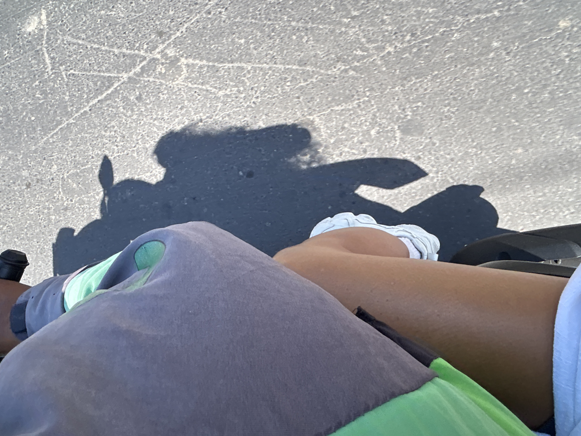 The image shows a close-up view of a person's legs and torso, viewed from a high angle. The person is wearing a grey sweatshirt with green accents, a black object partially visible under the sweatshirt, white sneakers, and possibly white shorts or underwear below the frame. The person's skin appears tanned and slightly dewy. They are sitting on something that casts a shadow, and the shadow of the person's head, complete with what appears to be a hat, is visible on the asphalt ground. The ground is grey, textured, and shows some faint white markings, possibly from chalk or paint. The lighting suggests it is daytime. The location is specified as North Kuta, Indonesia. The scene is likely outdoors, given the shadow and asphalt surface. The activity appears to be stationary or slowly moving, as indicated by the static nature of the shadow and the composition of the shot.