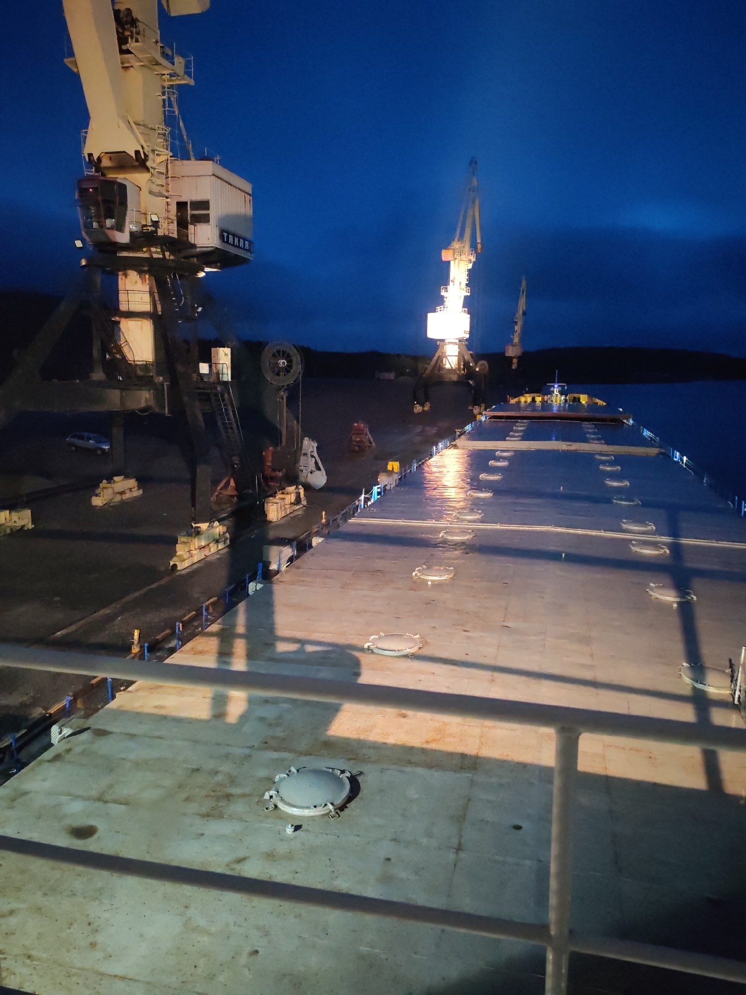 The image depicts a port scene at night. In the foreground, there is a deck with several round hatches. In the background, there are three large cranes; one with a visible "TARAK" inscription. The port is located in the city of Shalsky, Russia. The environment appears to be a working industrial area, with the cranes likely used for loading and unloading cargo. The sky is dark, illuminated by the lights of the cranes and the dock.